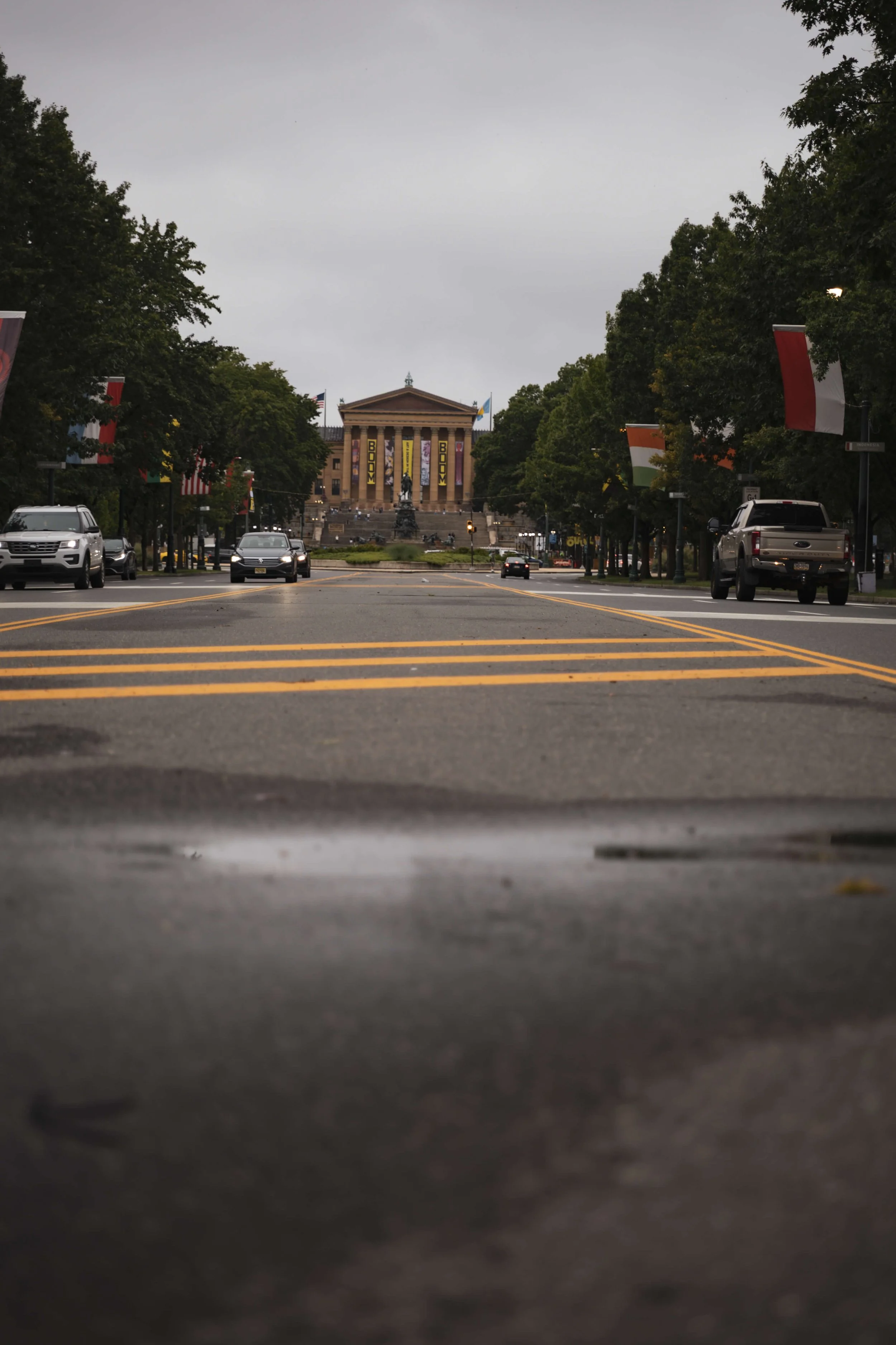 Street view with cars and traffic lines, leading to a large historic building with columns and statues, decorated with banners and flags.