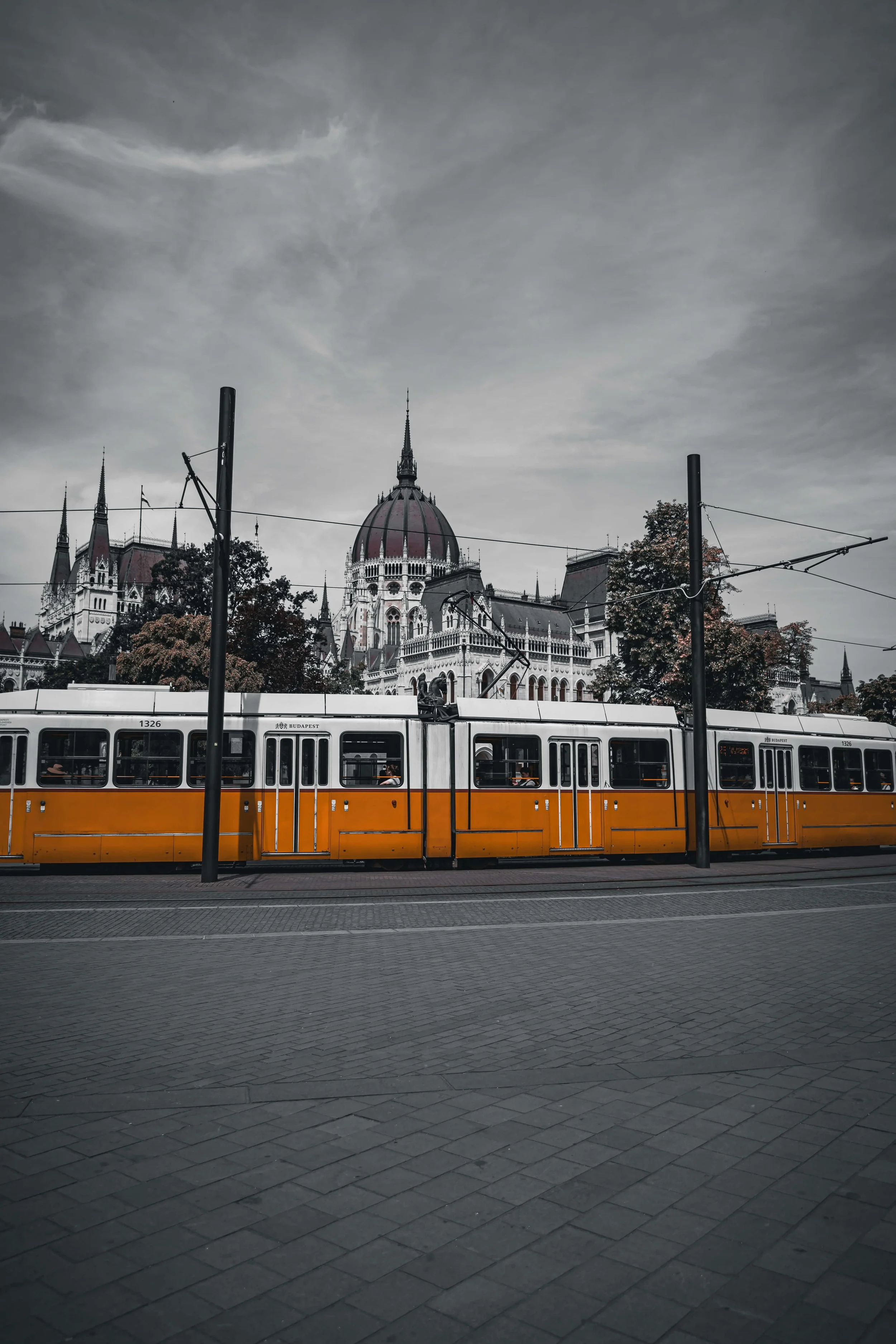Image of a yellow and white tram moving along tracks on a city street with a historic building with a domed roof and spire in the background, under a cloudy sky.