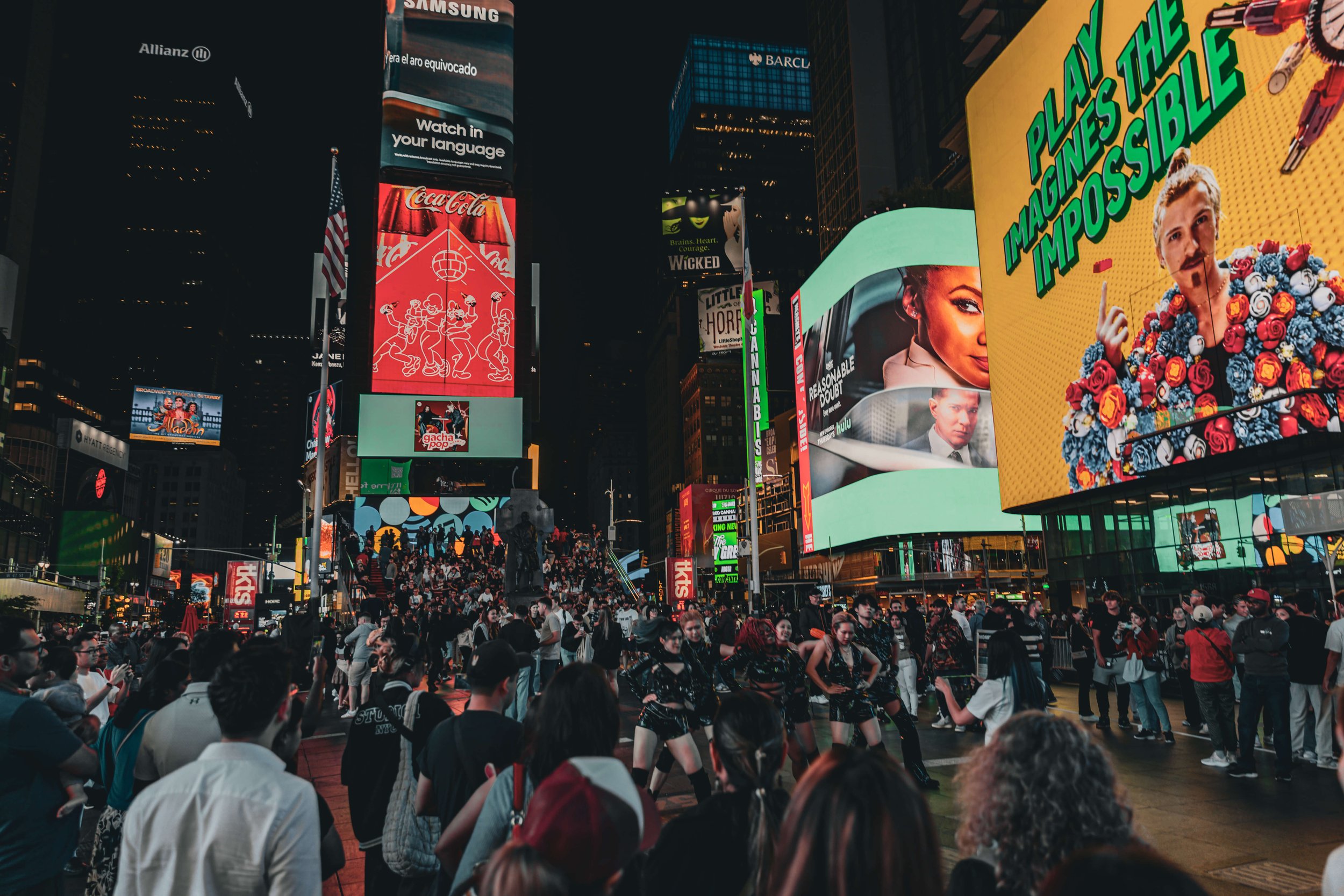 Crowd of people gathered in Times Square at night with bright electronic billboards and advertisements.
