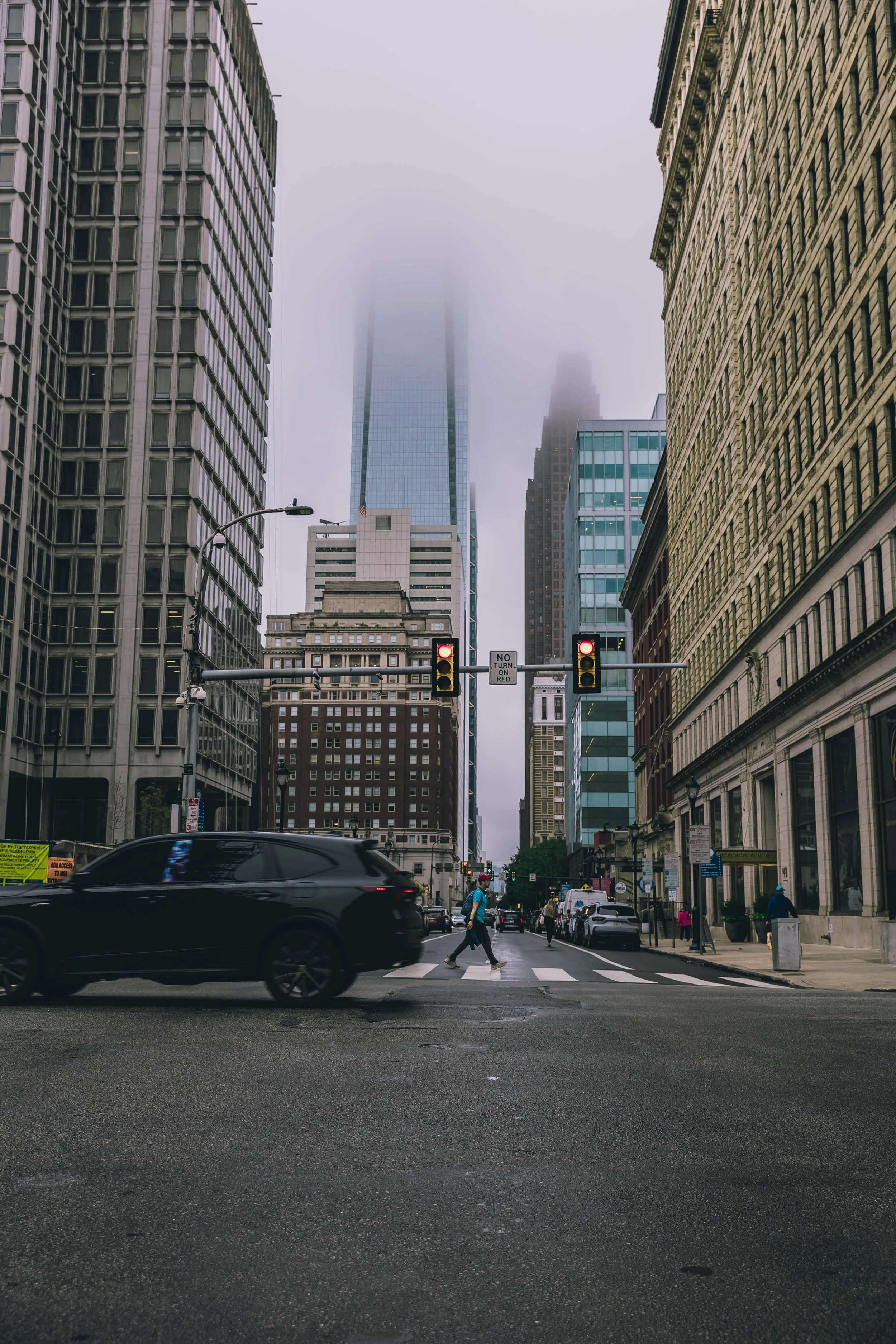 A city street scene with tall buildings, some partially obscured by fog. People are crossing at a crosswalk while a black vehicle passes by. Traffic lights are red, and there are pedestrians walking along the sidewalks in an urban environment.