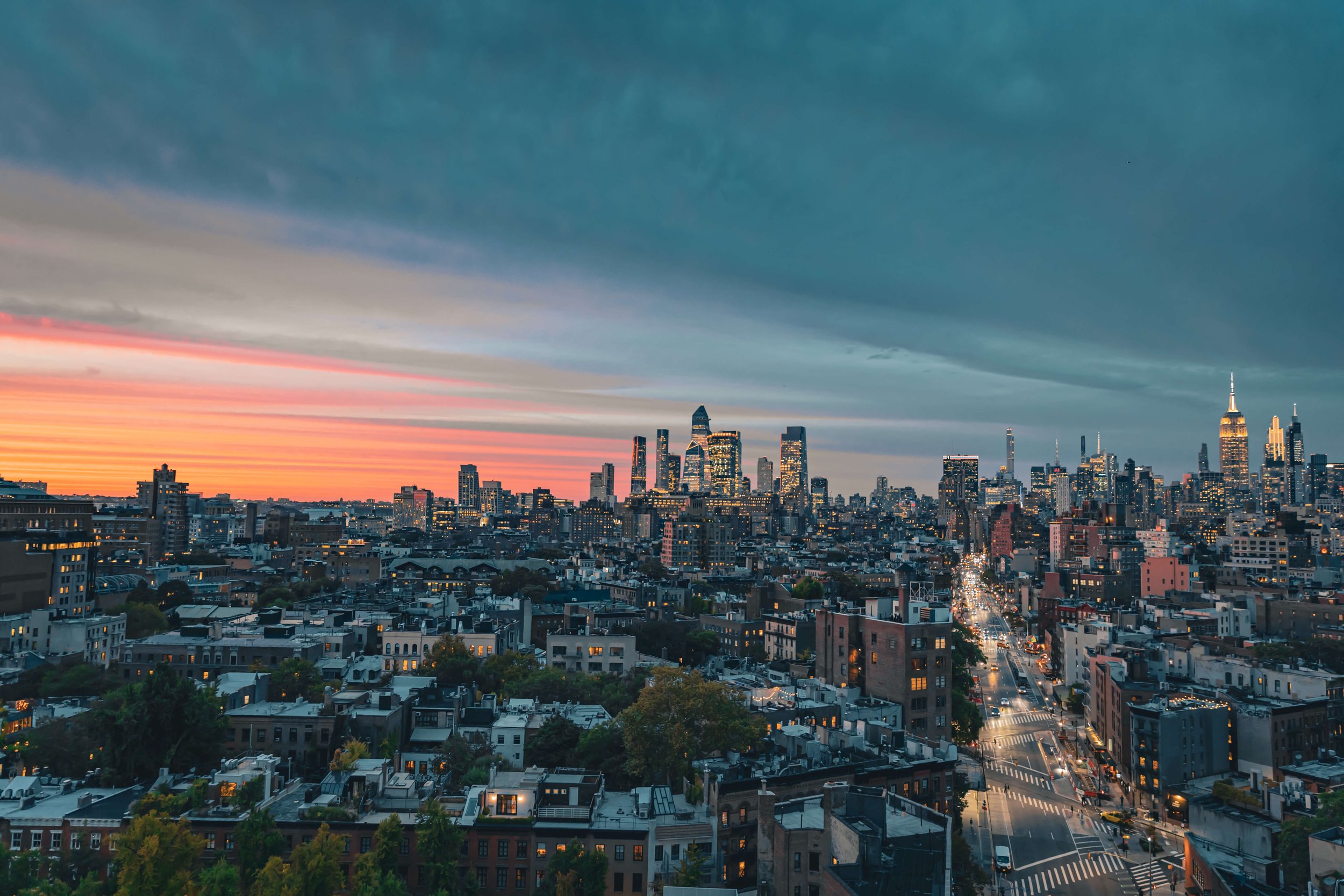A panoramic view of New York City skyline at dusk with a colorful sunset, tall skyscrapers including Empire State Building, and traffic on the streets.