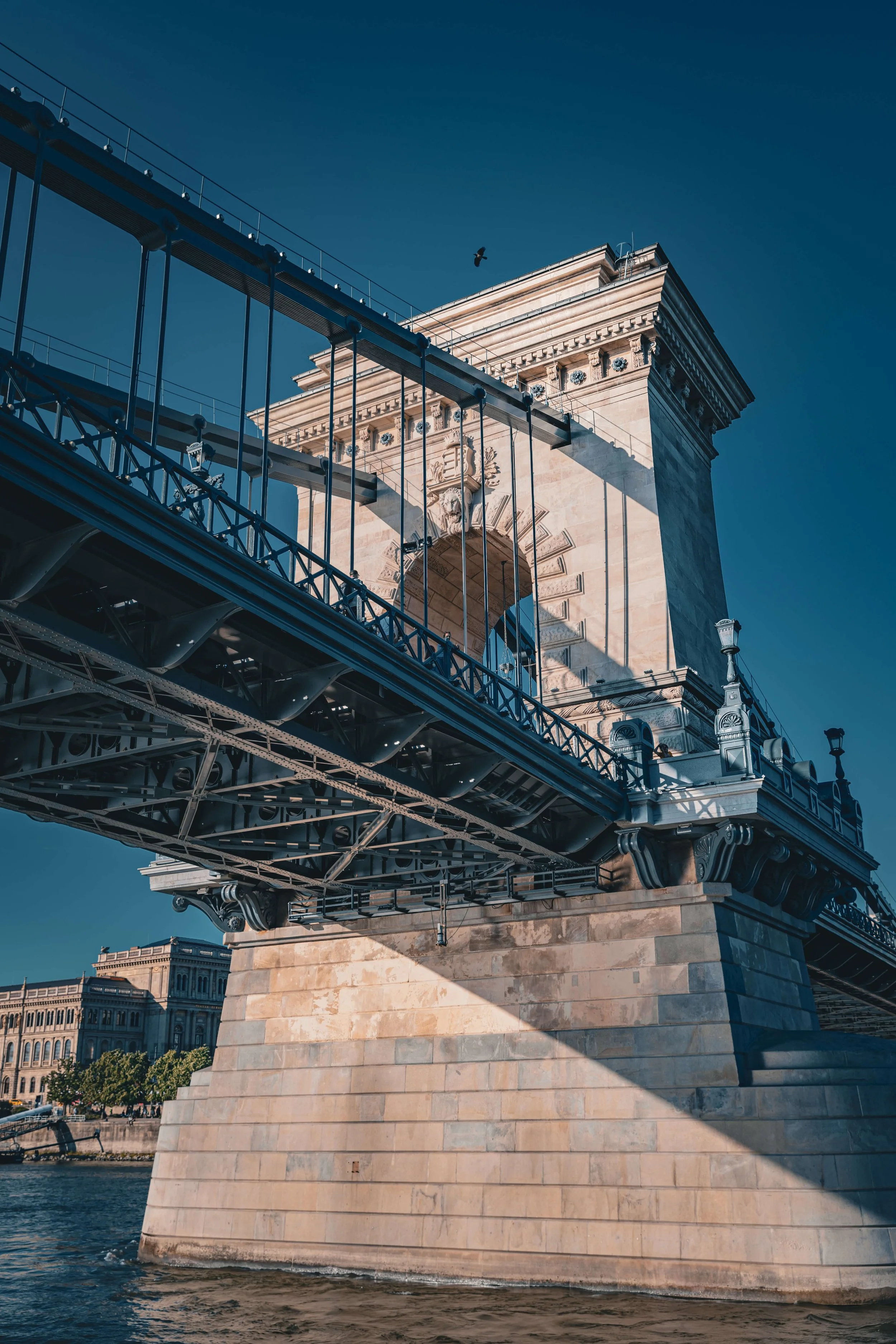Close-up view of the Chain Bridge in Budapest, Hungary, showing its stone tower and the suspension cables extending from it, with a bird flying in the clear blue sky above.