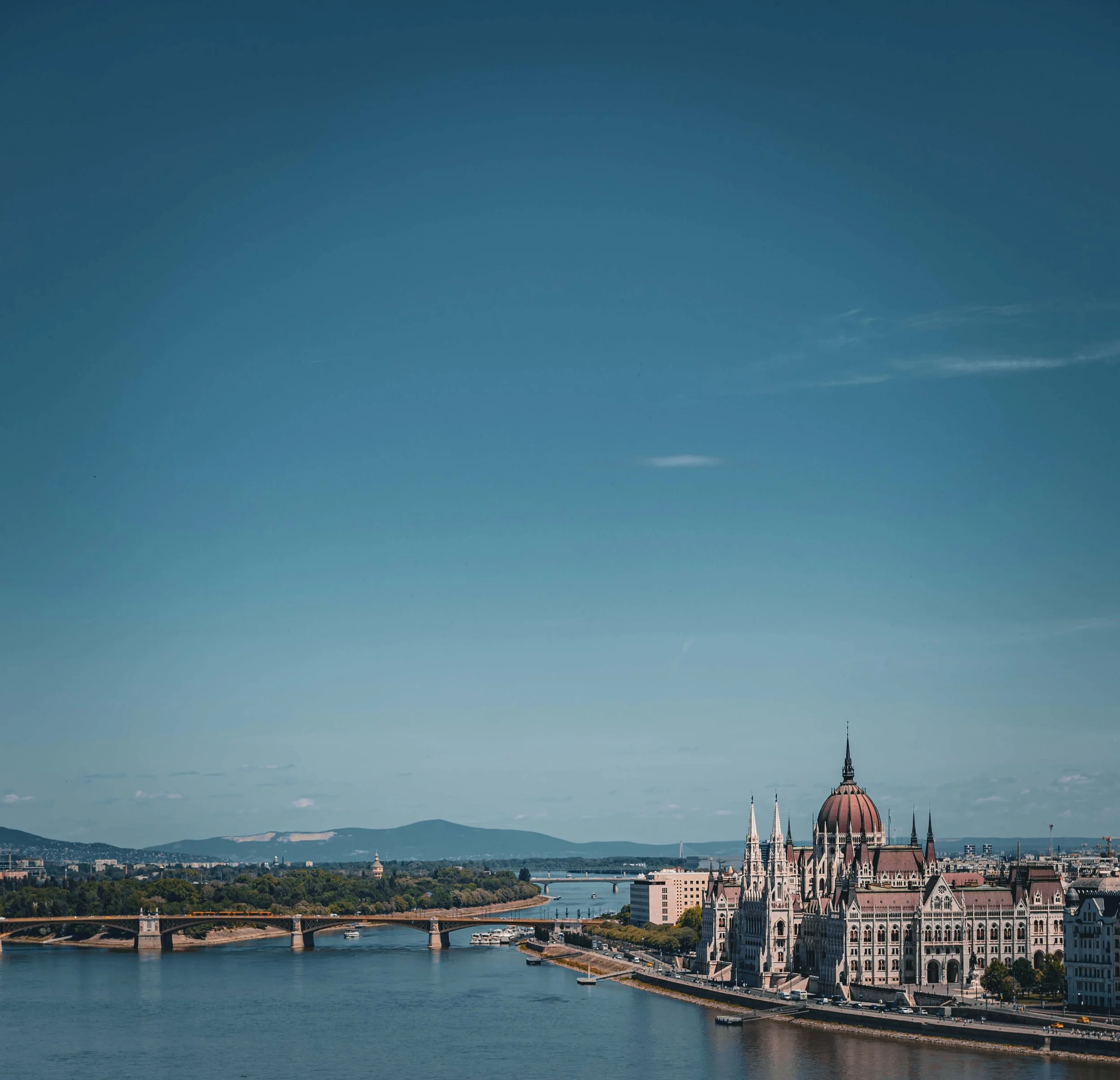 A panoramic cityscape featuring a river with boats, bridges, and historic buildings, with a prominent church spire and a large dome in the background, under a clear blue sky with few clouds.