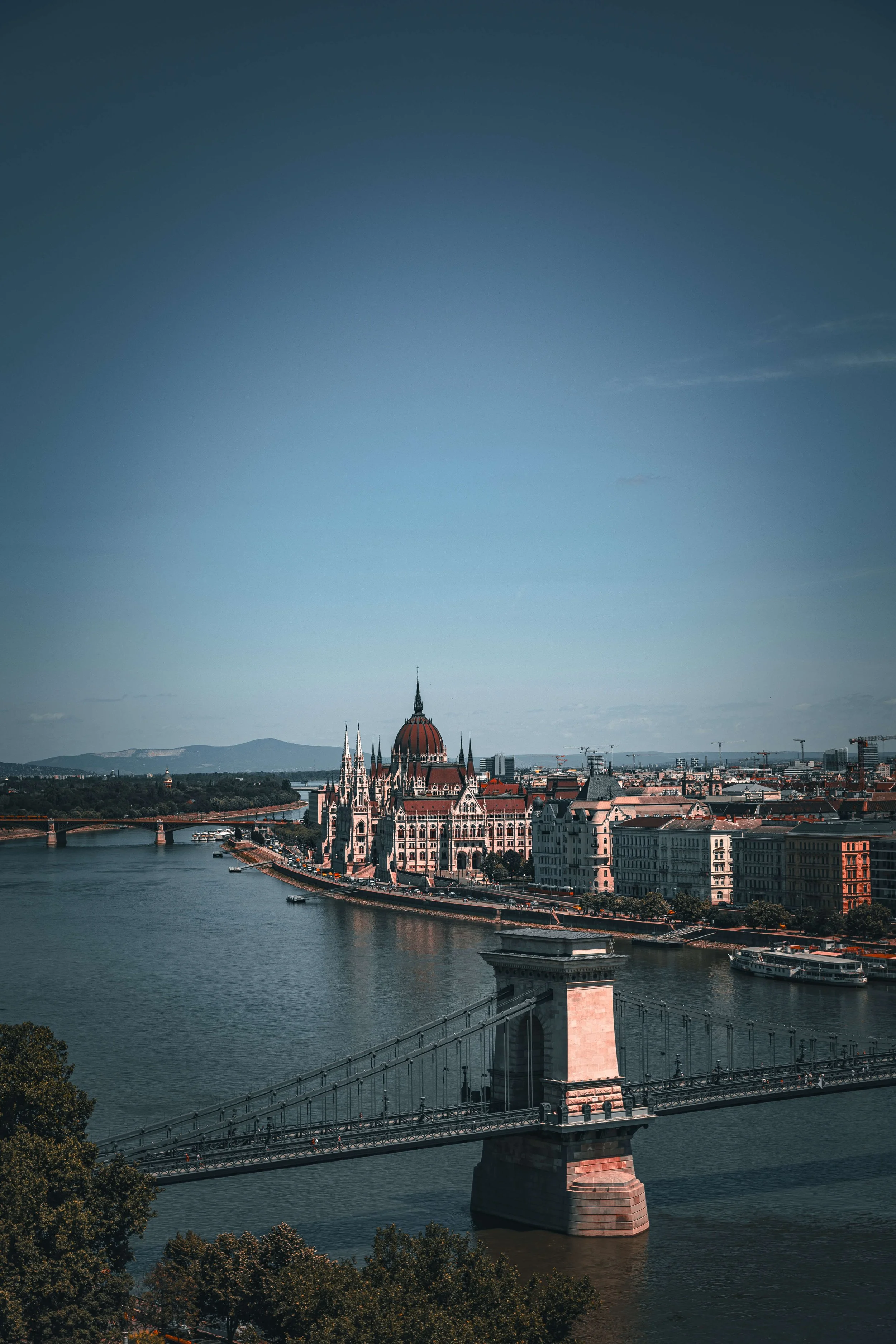 A cityscape featuring the Hungarian Parliament Building in Budapest, situated along the Danube River, with a bridge crossing the river in the foreground and mountains in the background under a partly cloudy sky.