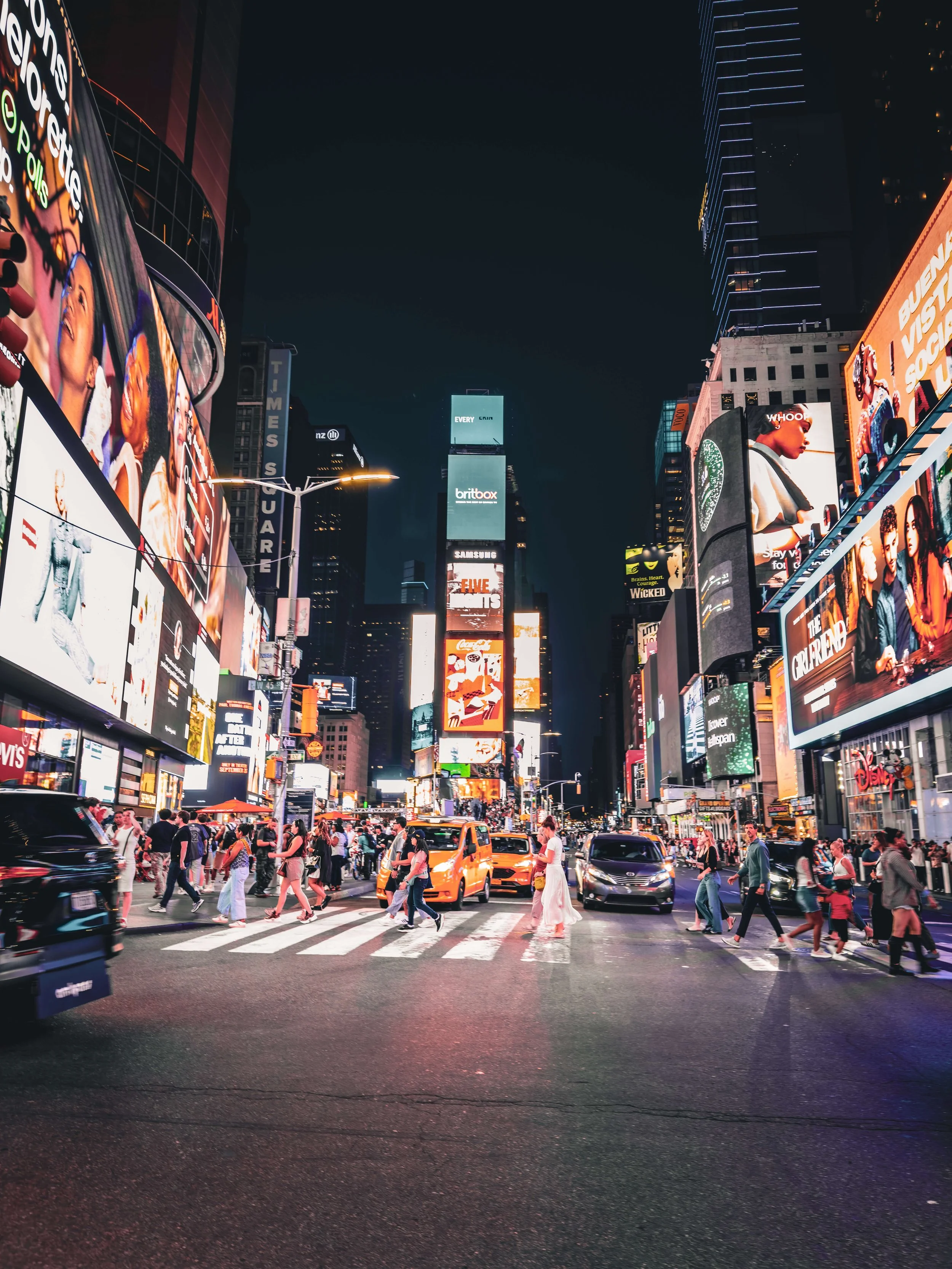 Nighttime scene in Times Square, New York City, with bright electronic billboards, tall buildings, and a crowd of pedestrians crossing the street among yellow taxis and other vehicles.