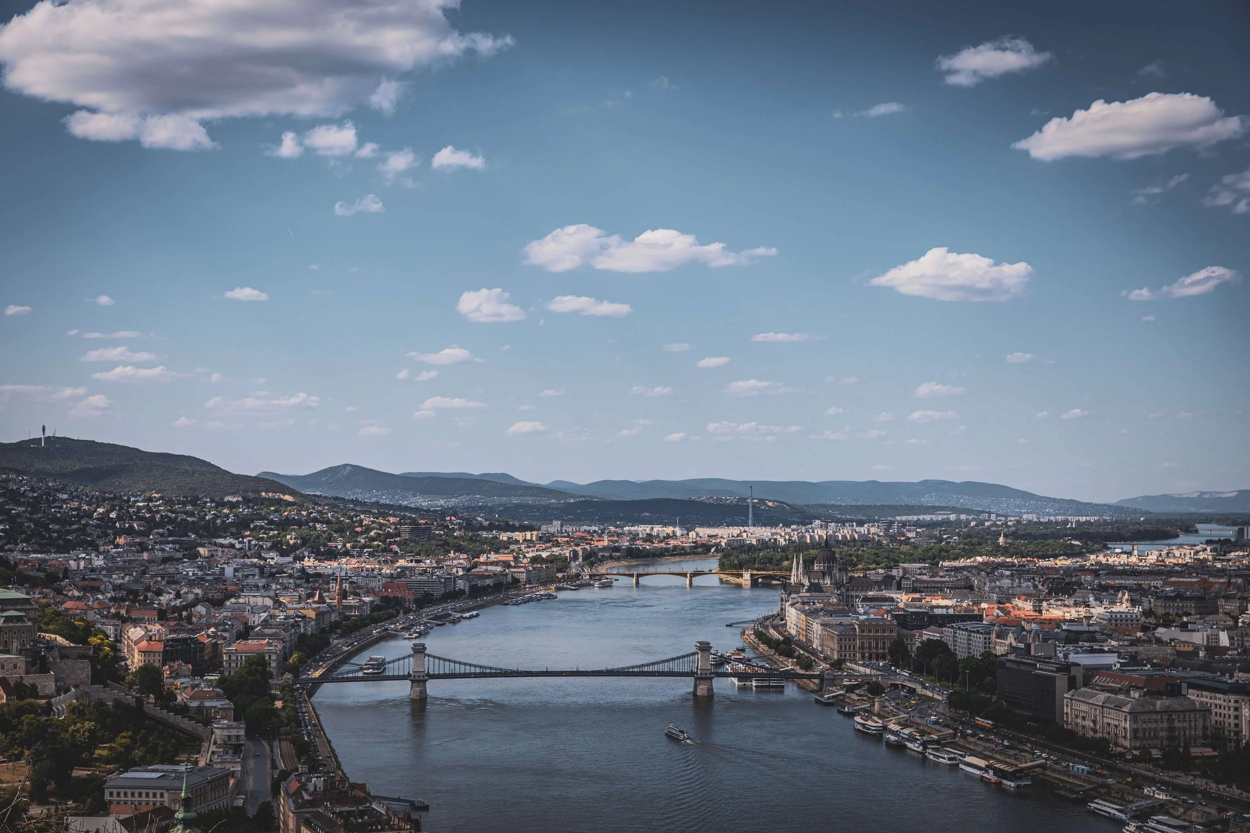 A panoramic view of a city with a river running through it, multiple bridges crossing the river, and a large hill in the background under a partly cloudy sky.