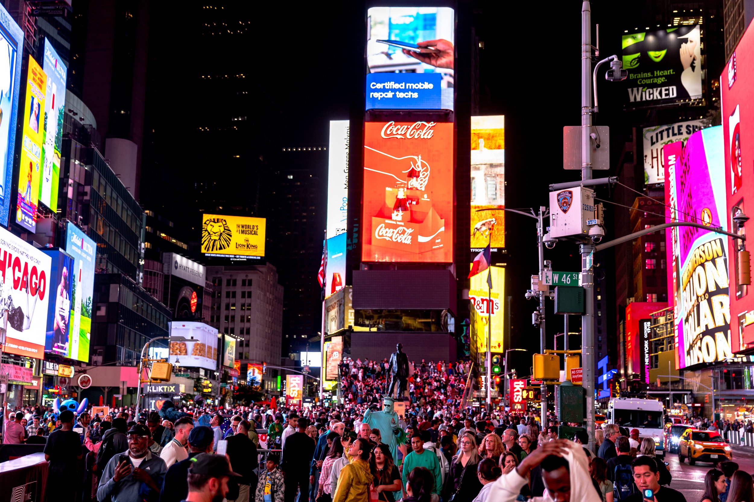Brightly lit Times Square at night with digital billboards, a large crowd of people, and taxis on the street.