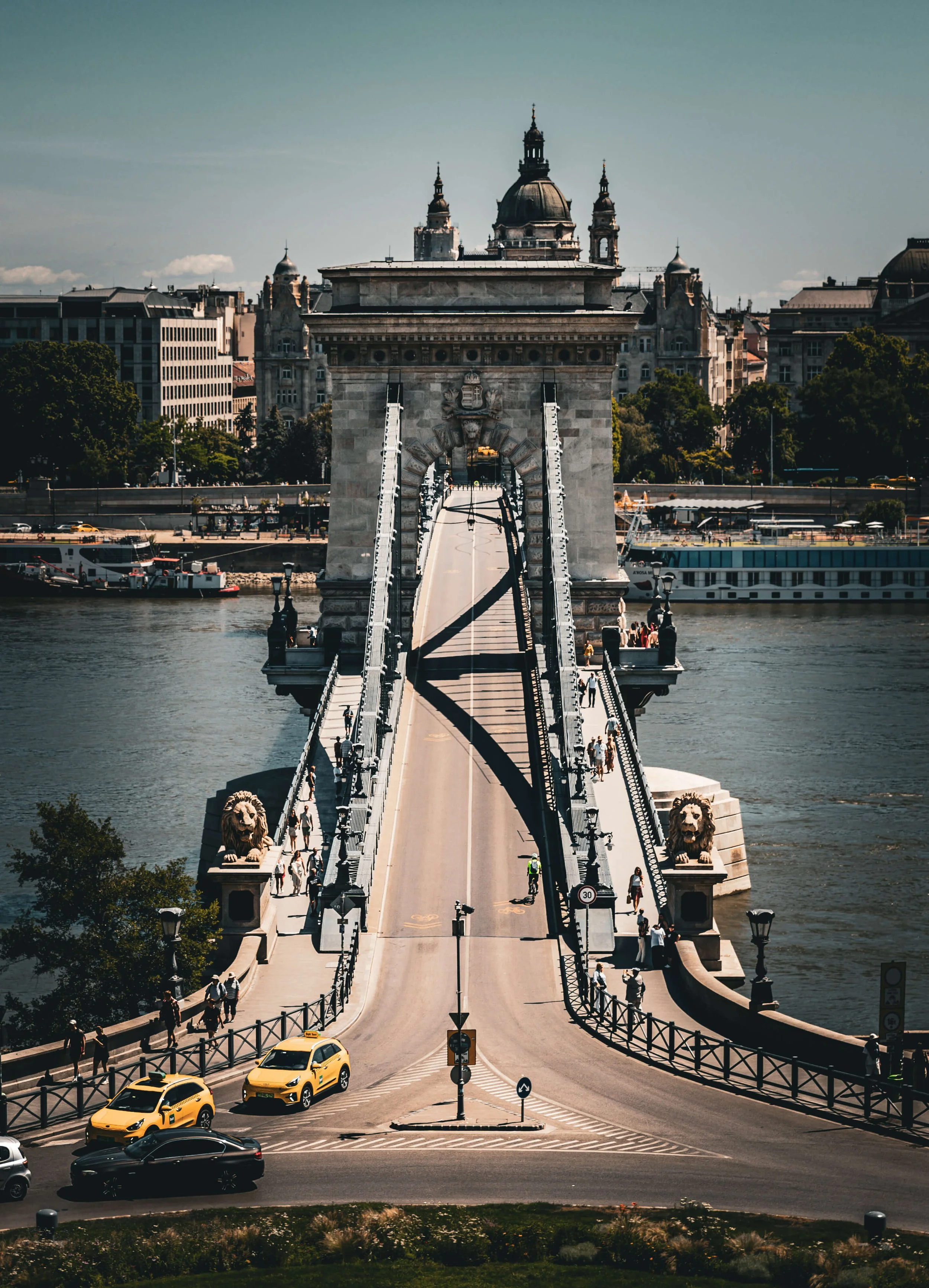The Chain Bridge in Budapest, Hungary, viewed from the Pest side, with yellow taxis and cars crossing the bridge and historic buildings in the background.
