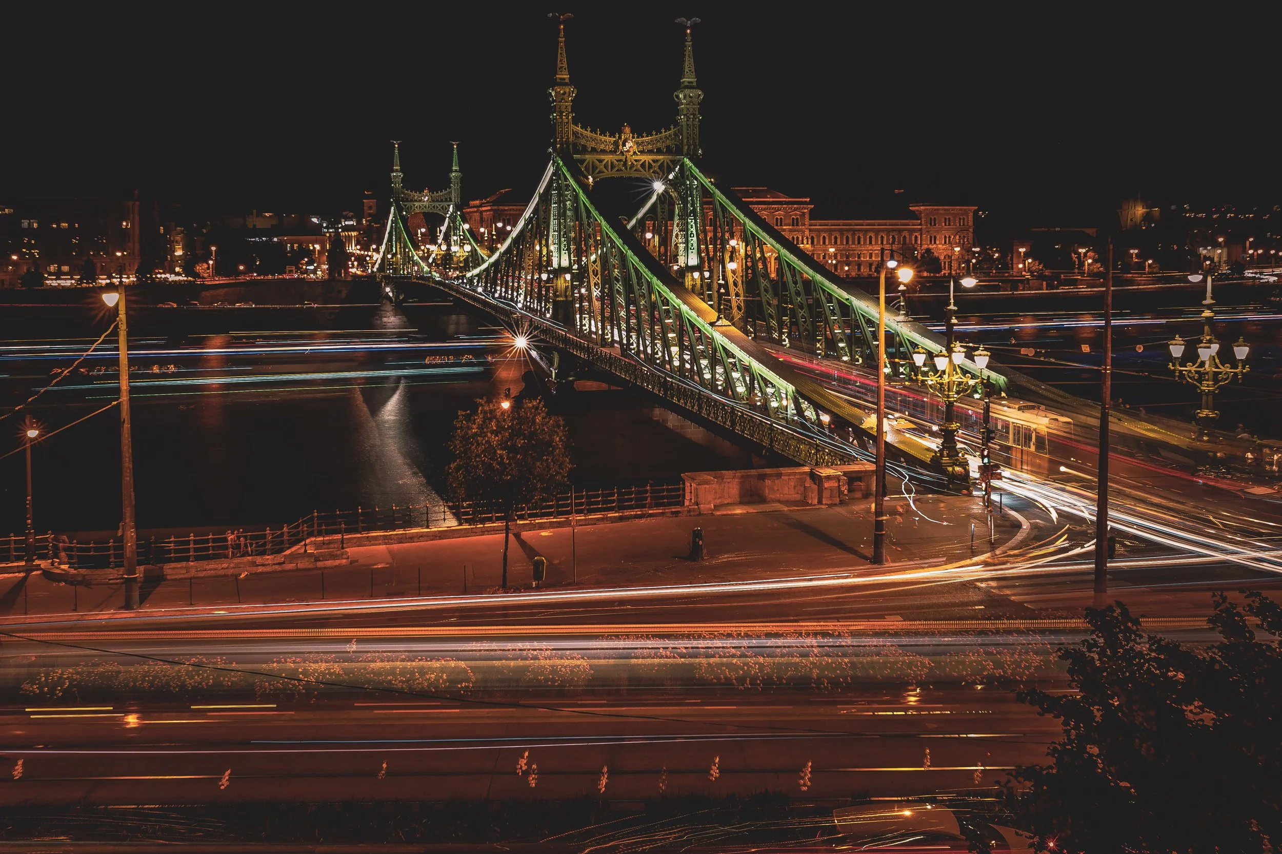Night view of the Liberty Bridge in Budapest, Hungary, illuminated with green lights, crossing the Danube River with city buildings in the background.