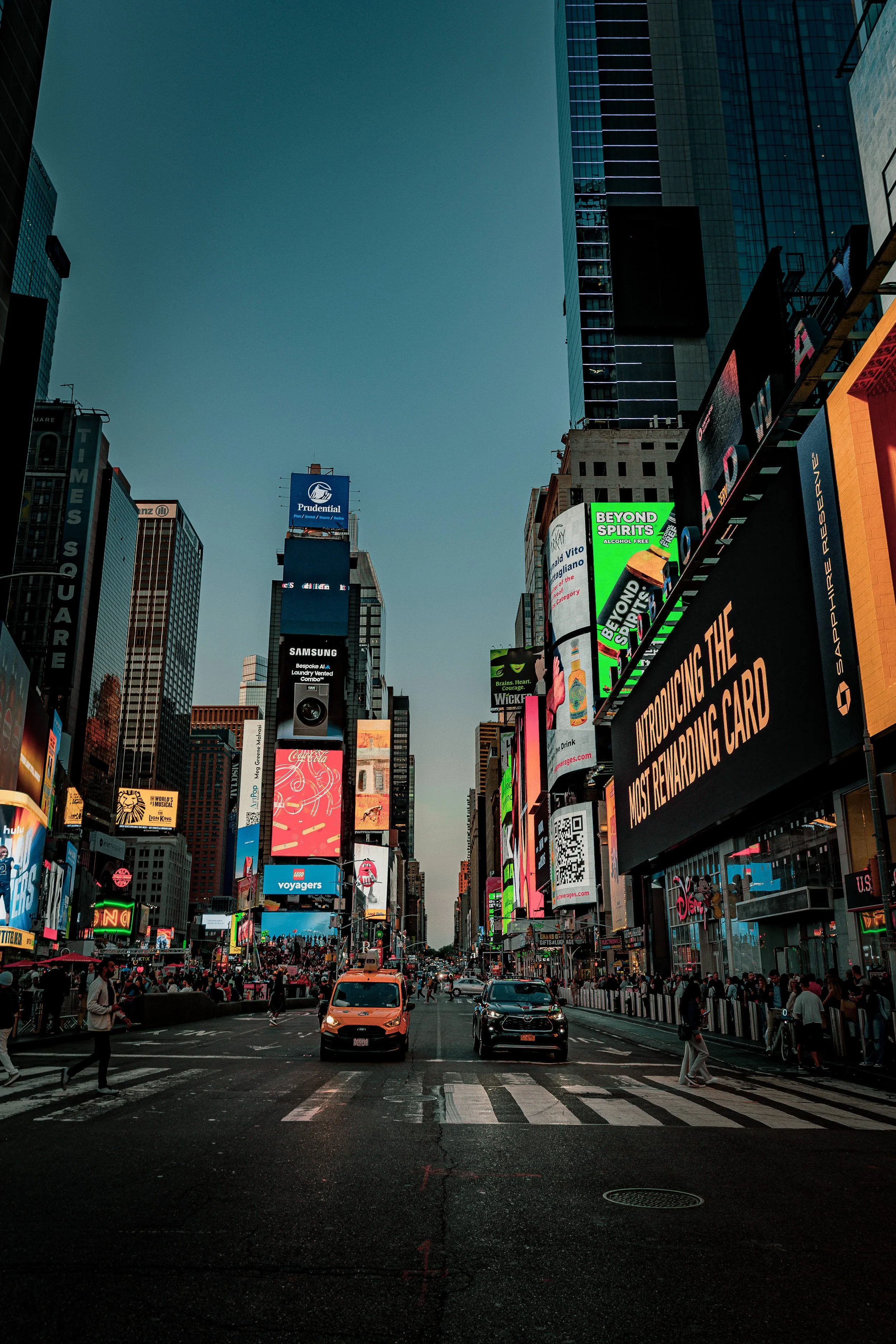 Times Square in New York City with tall buildings, digital billboards, and pedestrians crossing the street at dusk.
