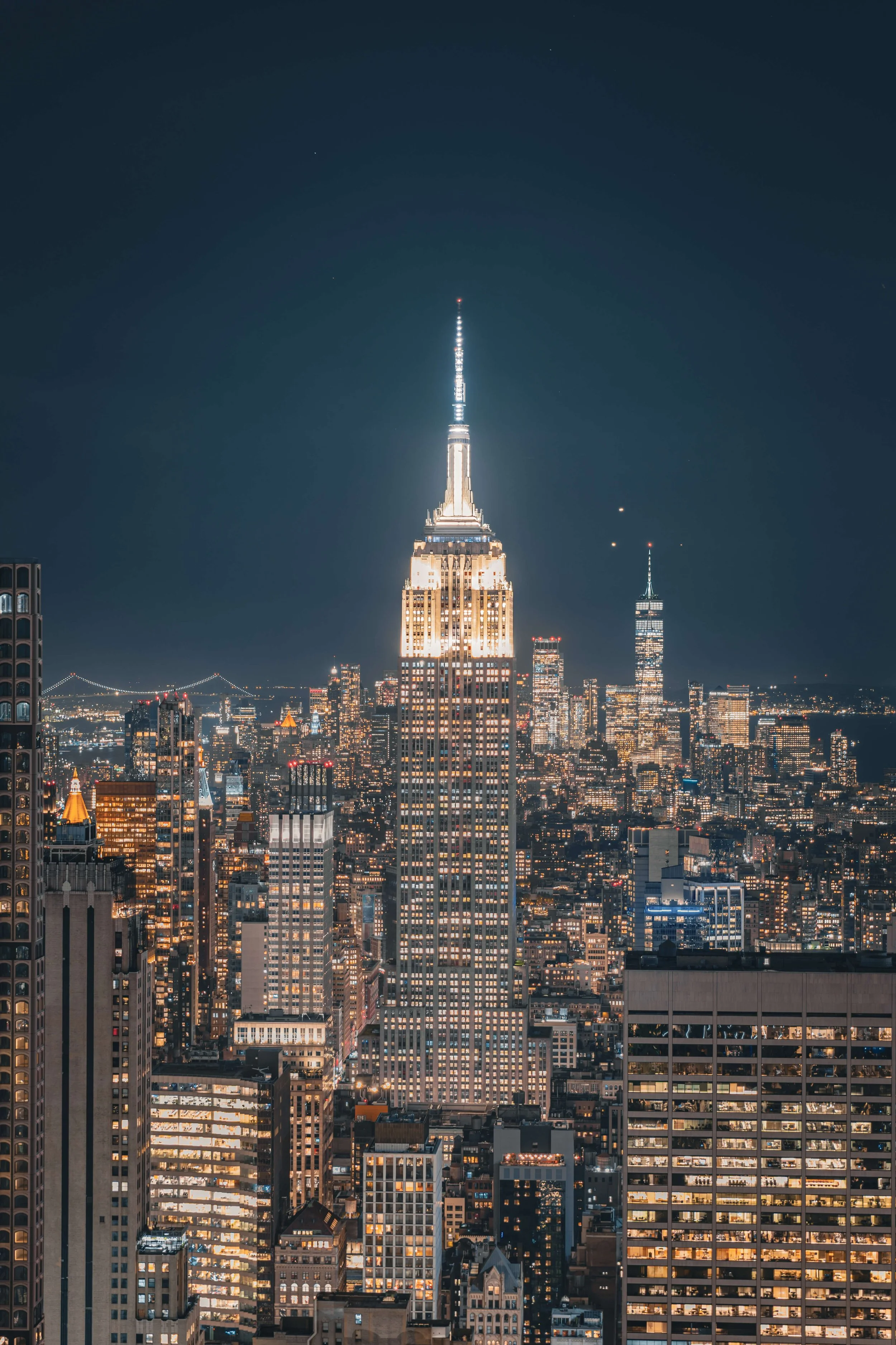 Nighttime view of the illuminated Empire State Building in New York City with surrounding skyscrapers and city lights.