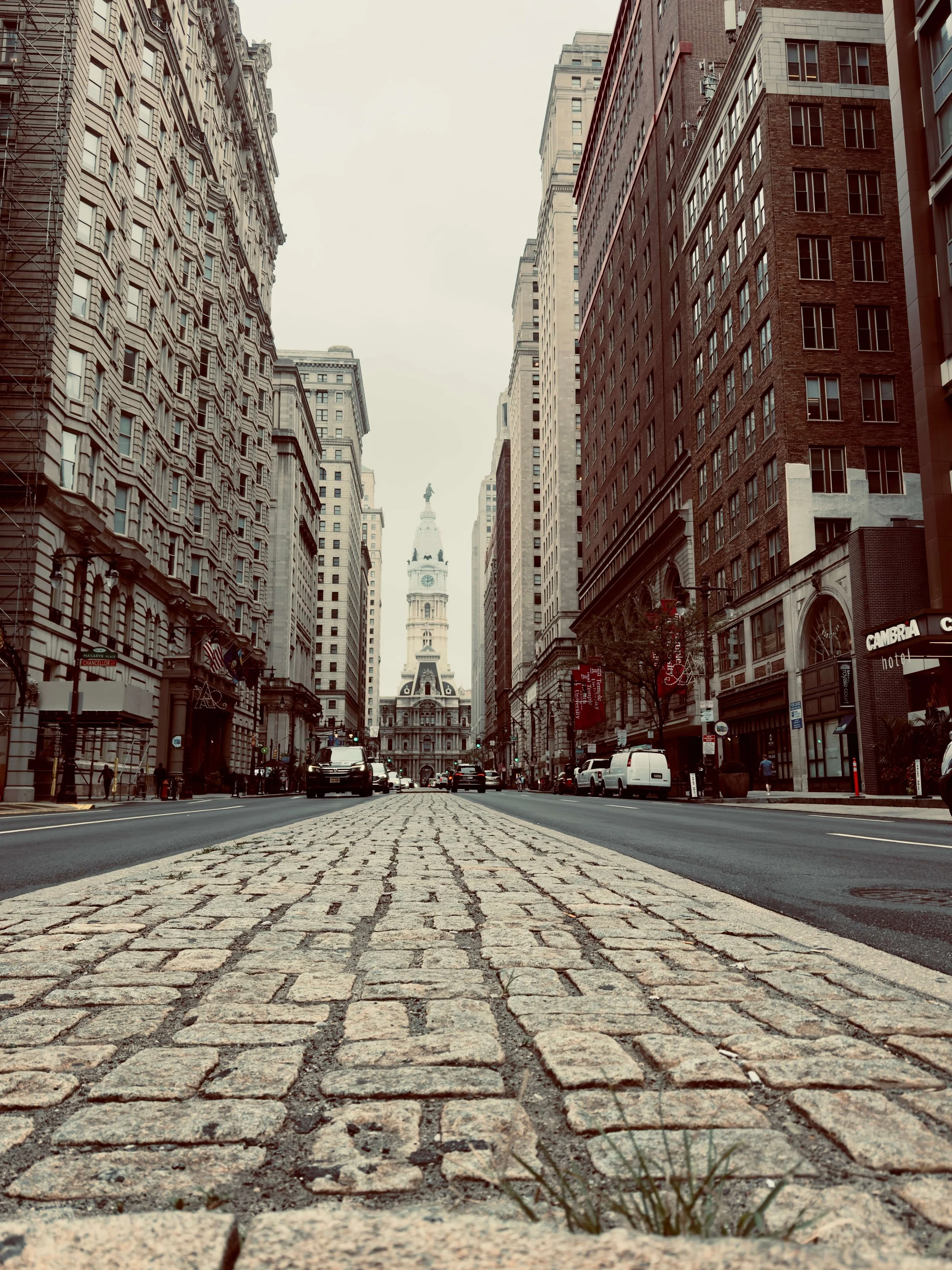 A city street with cobblestone pavement, tall buildings on both sides, and City Hall in the distance with its clock tower and statue on top.