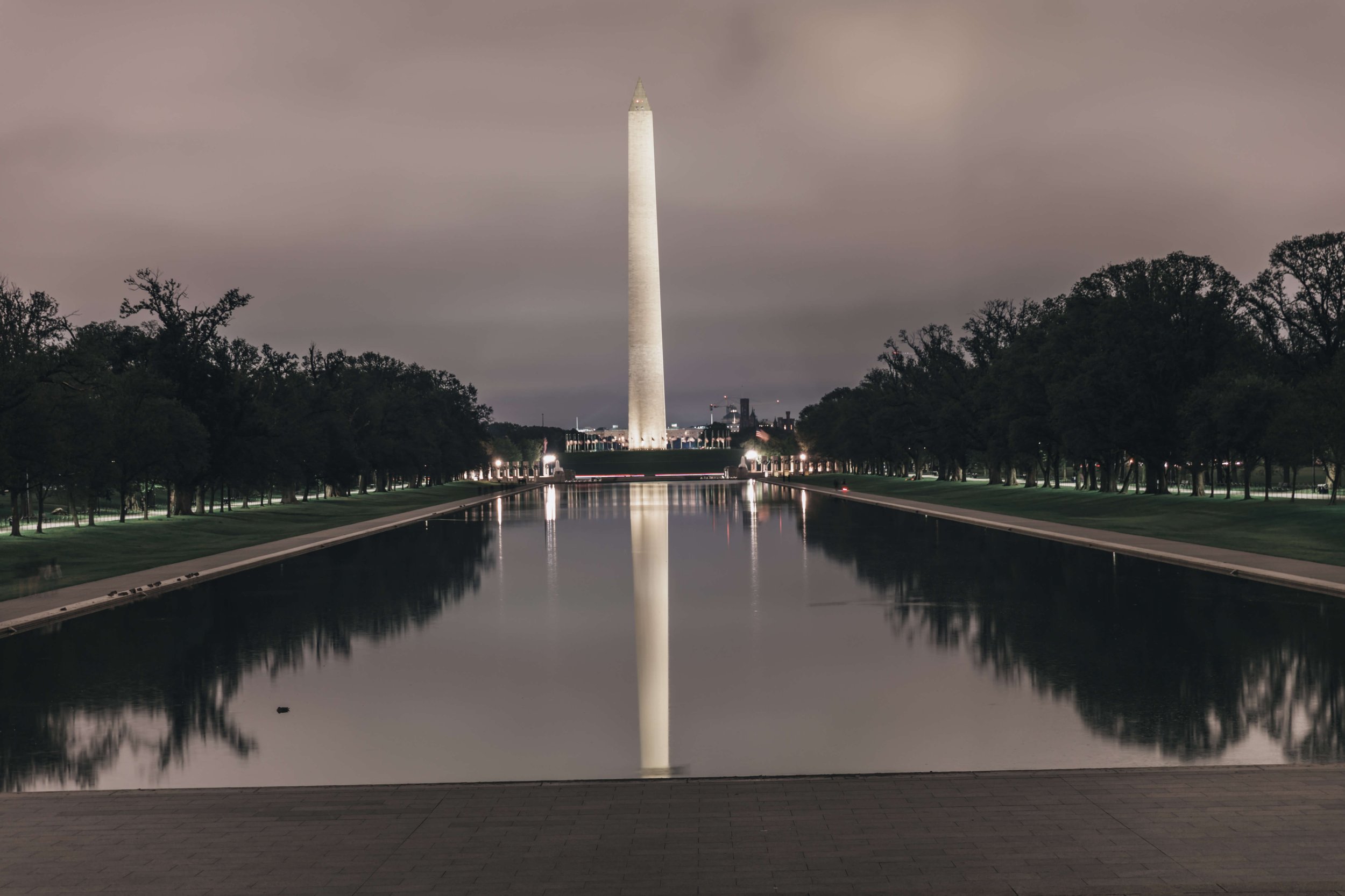 Night view of the Washington Monument reflected in the Reflecting Pool, with trees lining both sides and a cloudy sky overhead.