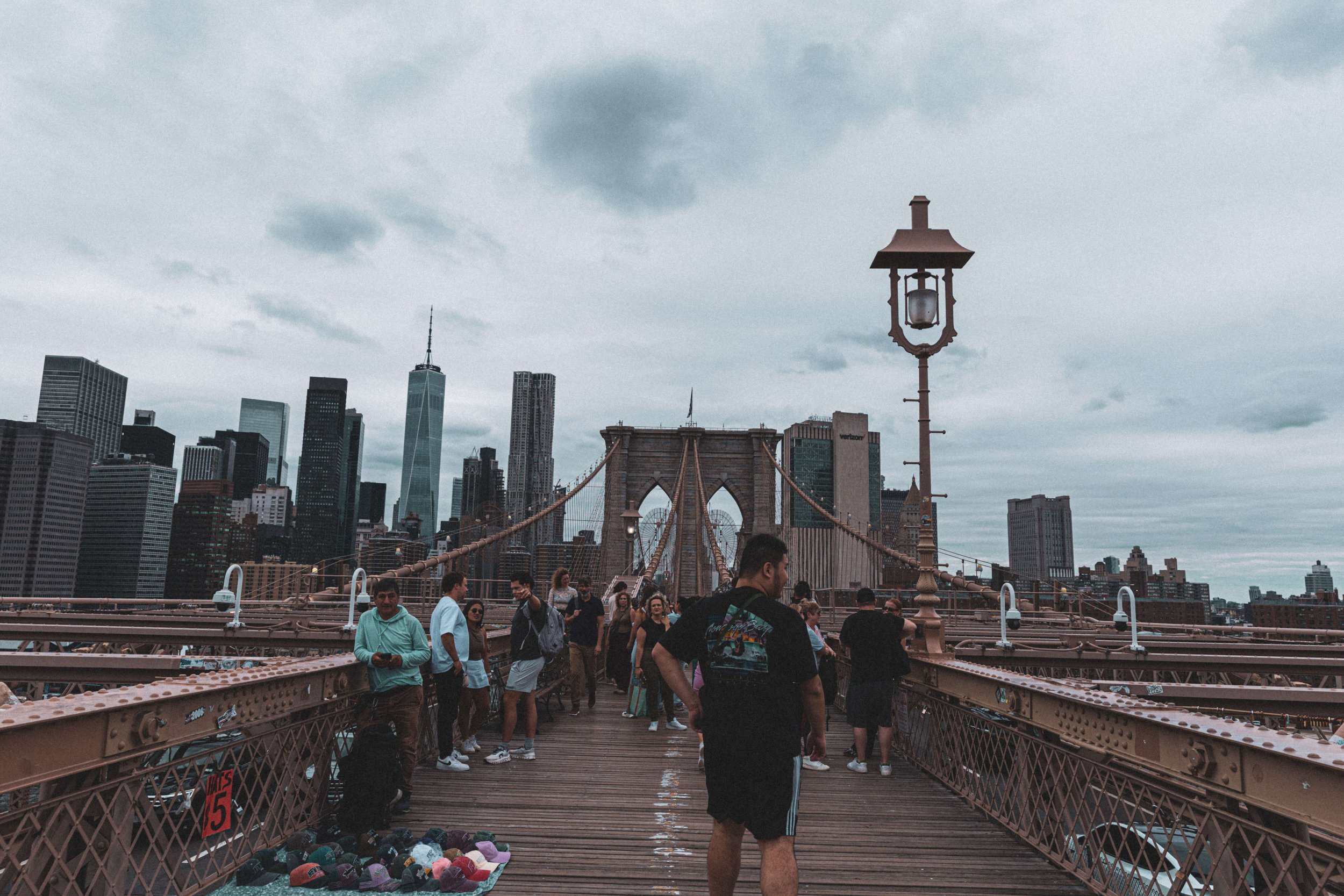 People walking on the Brooklyn Bridge in New York City with the Manhattan skyline in the background, overcast sky.