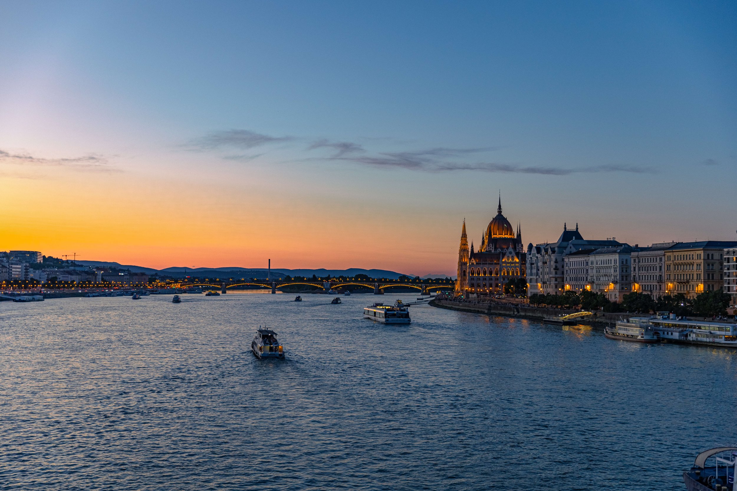 Sunset over the Danube River in Budapest, Hungary, with boats on the water and the Hungarian Parliament Building illuminated along the riverside.