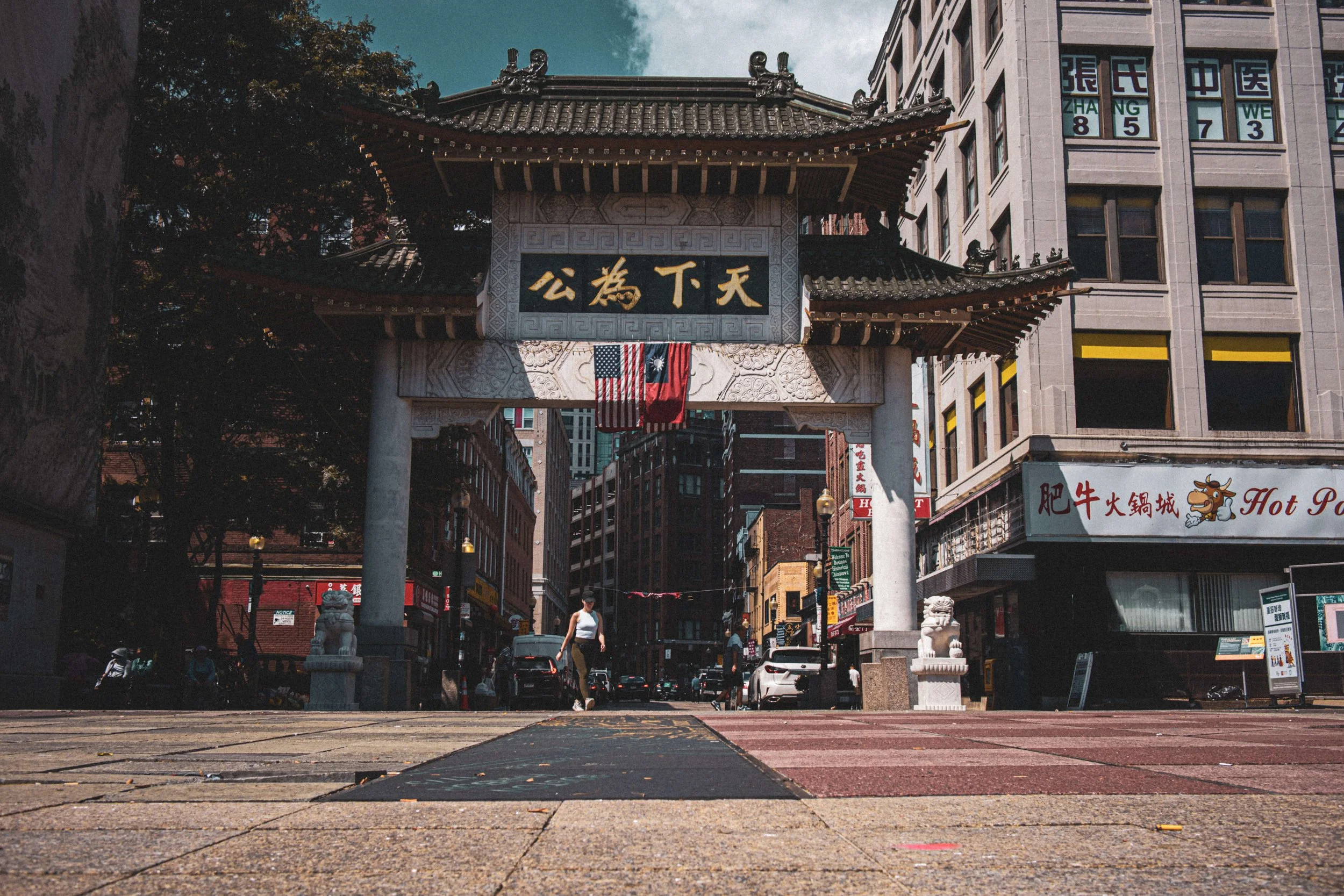 A traditional Chinese gate with Chinese characters and flags, leading into a busy urban street with buildings, shops, and pedestrians.