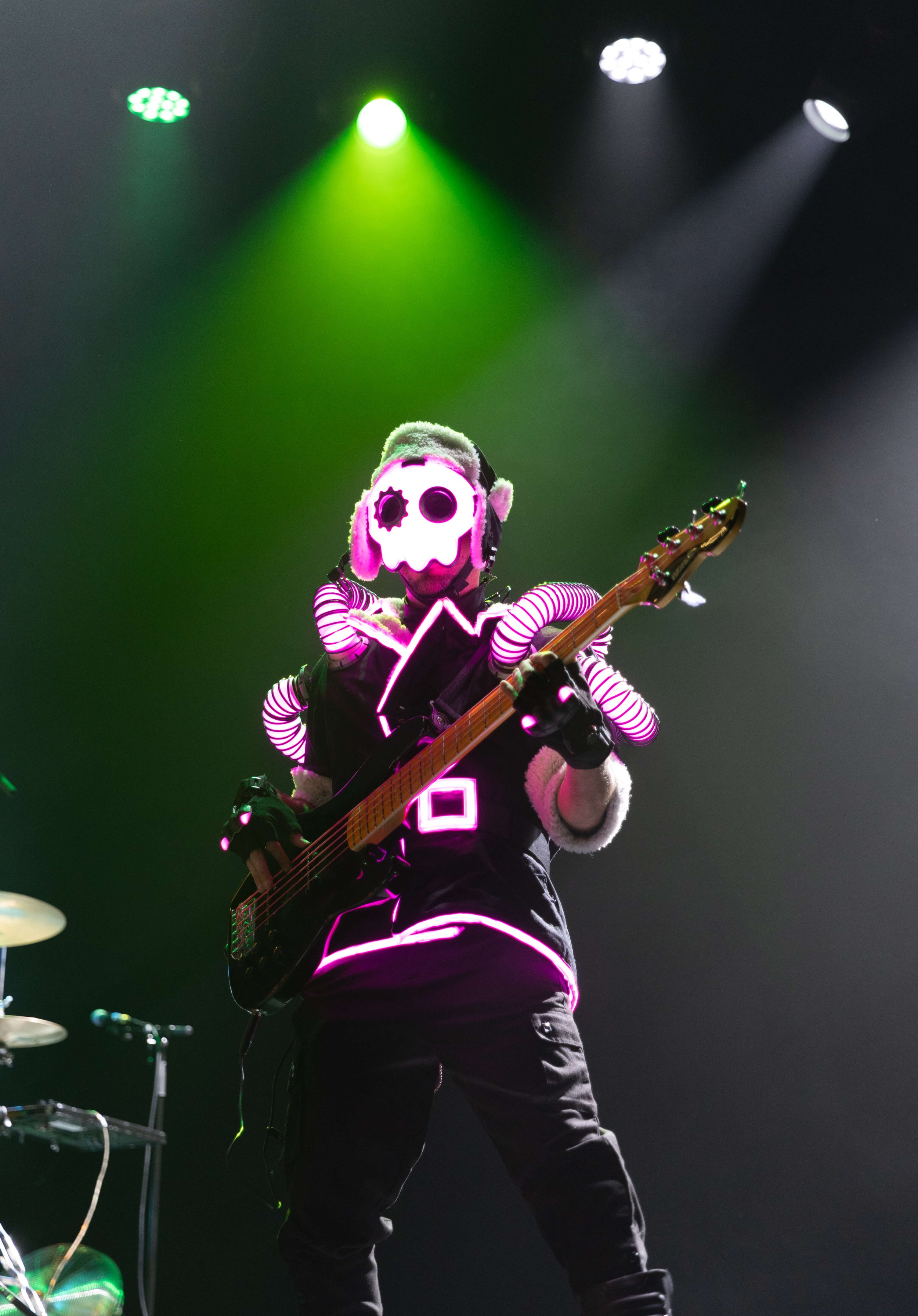 Musician on stage wearing a spooky skull mask with glowing pink and white lights, playing an electric guitar, illuminated by green and white stage lights.