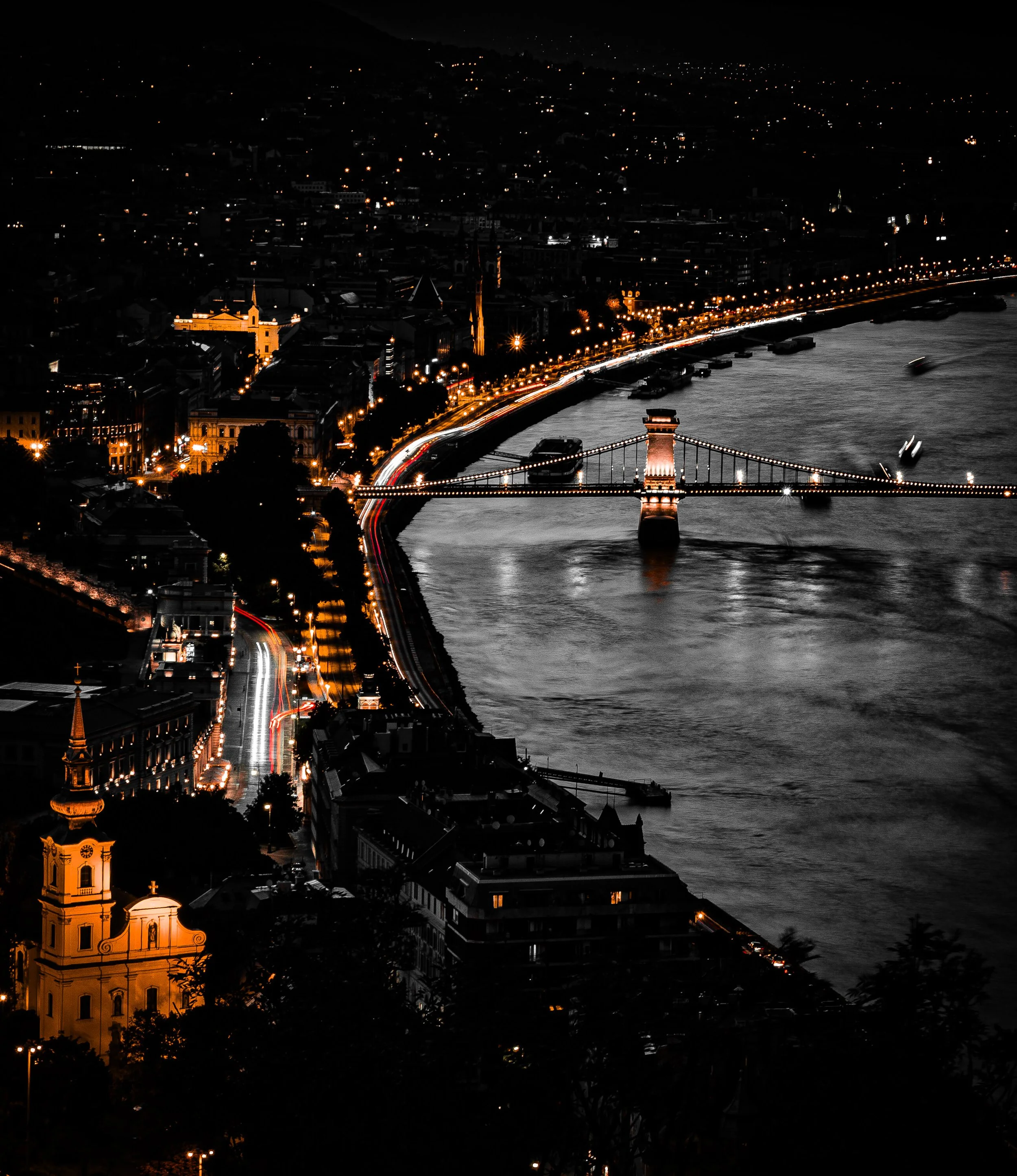 Nighttime aerial photograph of a city with illuminated buildings, a bridge over a river, and a hilly landscape in the background.