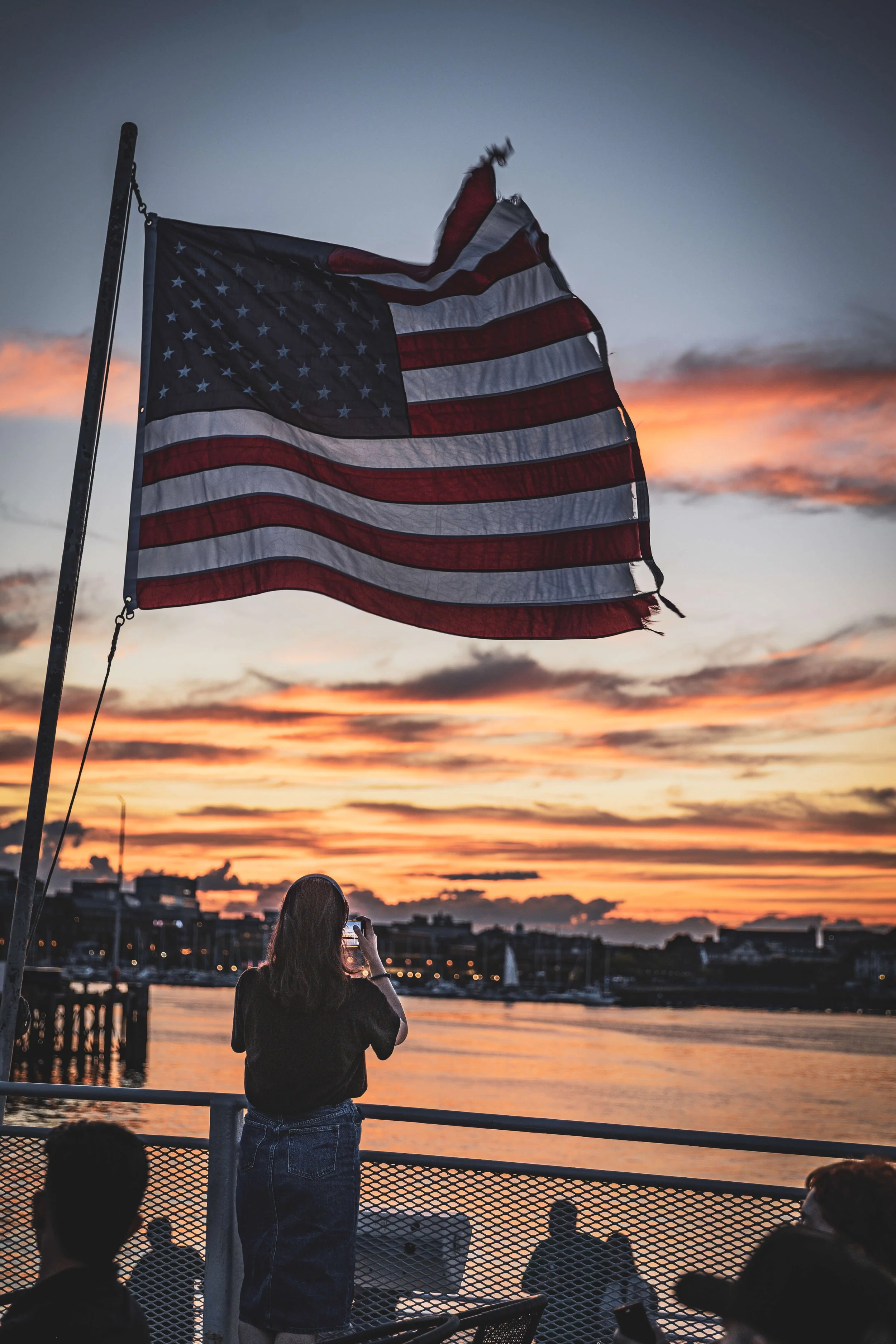 A faded American flag flying on a pole at sunset, with a woman taking a photo of the flag and river with buildings in the background.