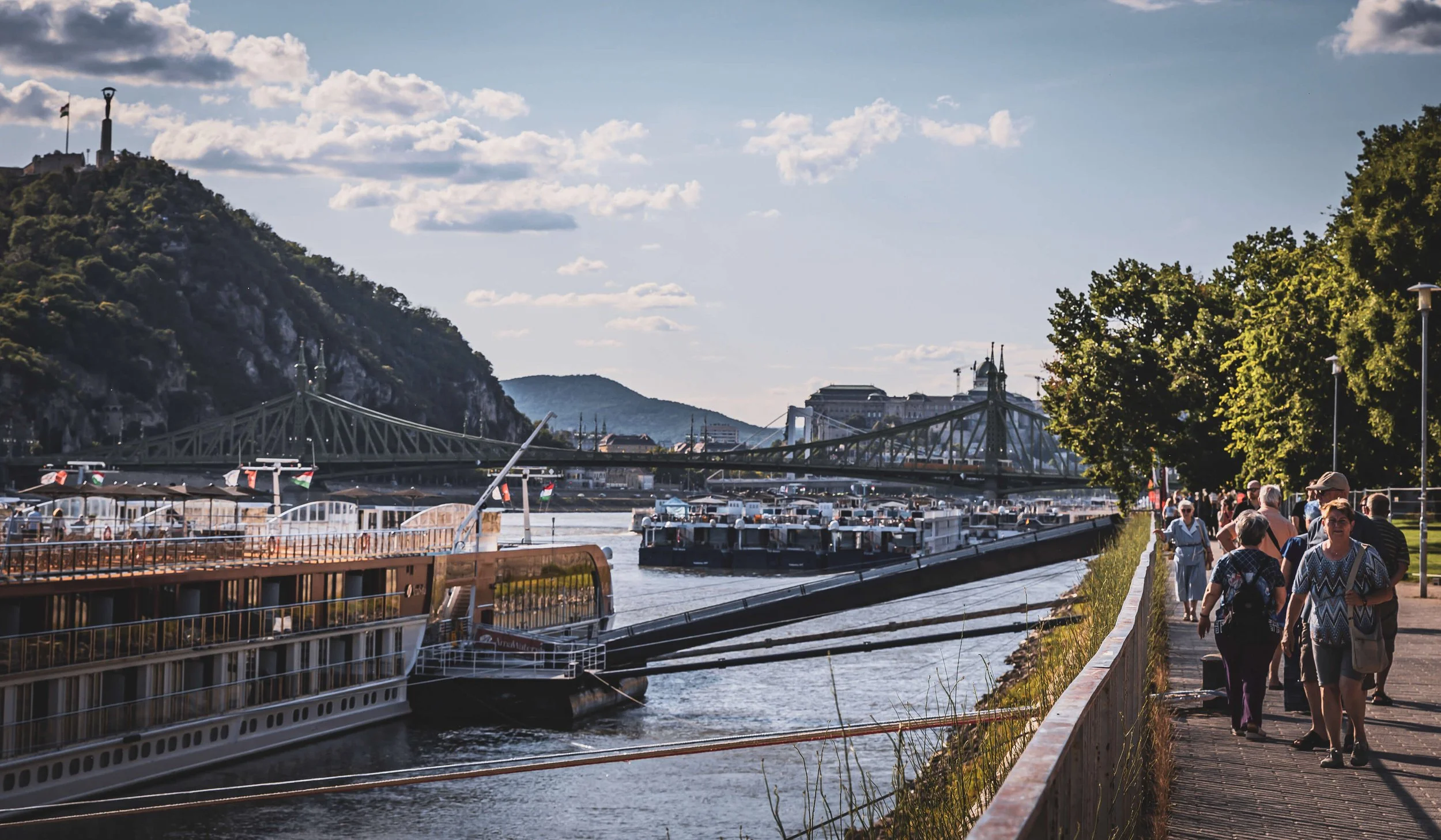 People walking along a riverside walkway with boats docked in the river, a bridge crossing the river, green trees, hills in the background, and a partly cloudy sky.