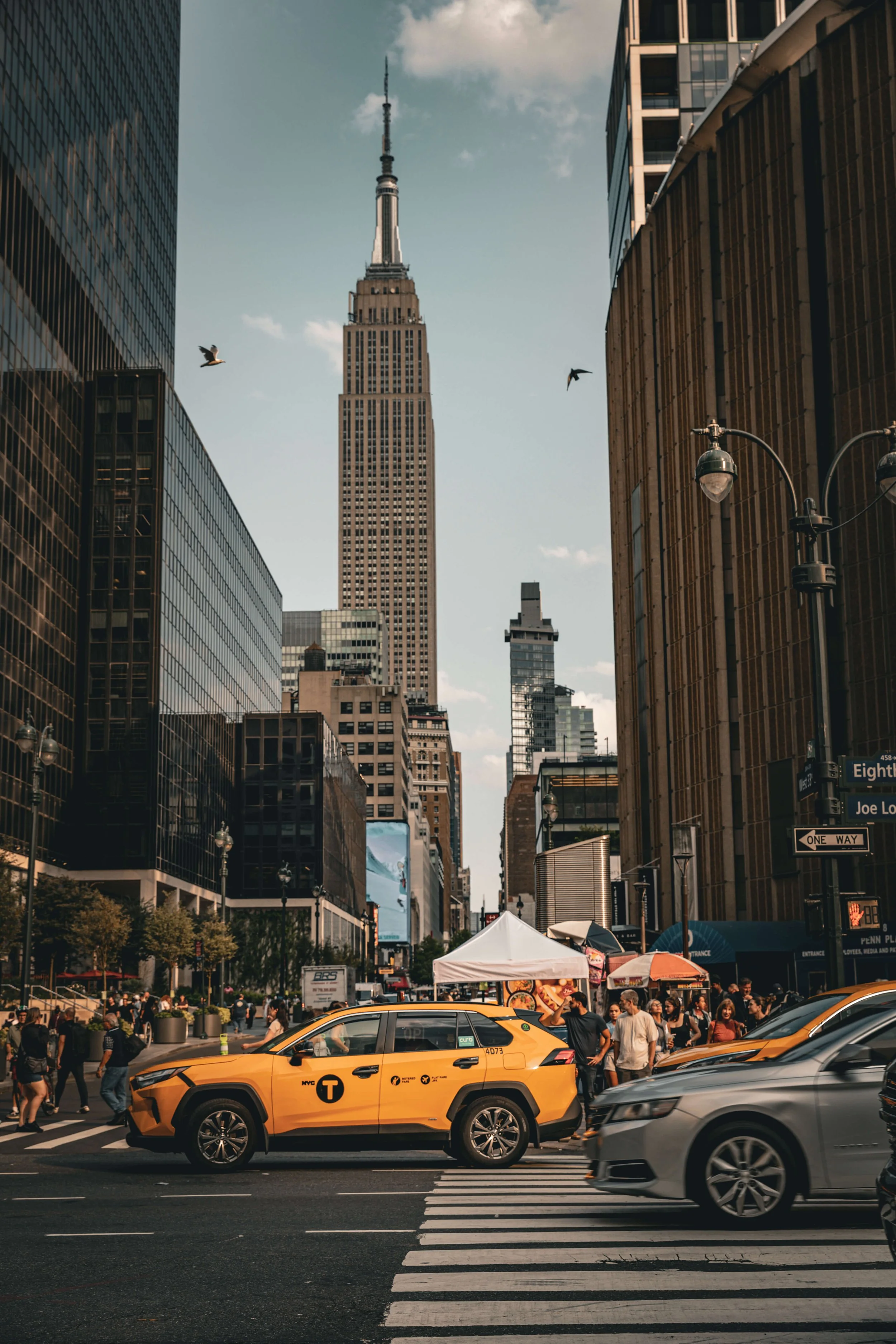 Street scene in New York City with the Empire State Building in the background, yellow taxi, pedestrians, storefronts, tents, and street signs.