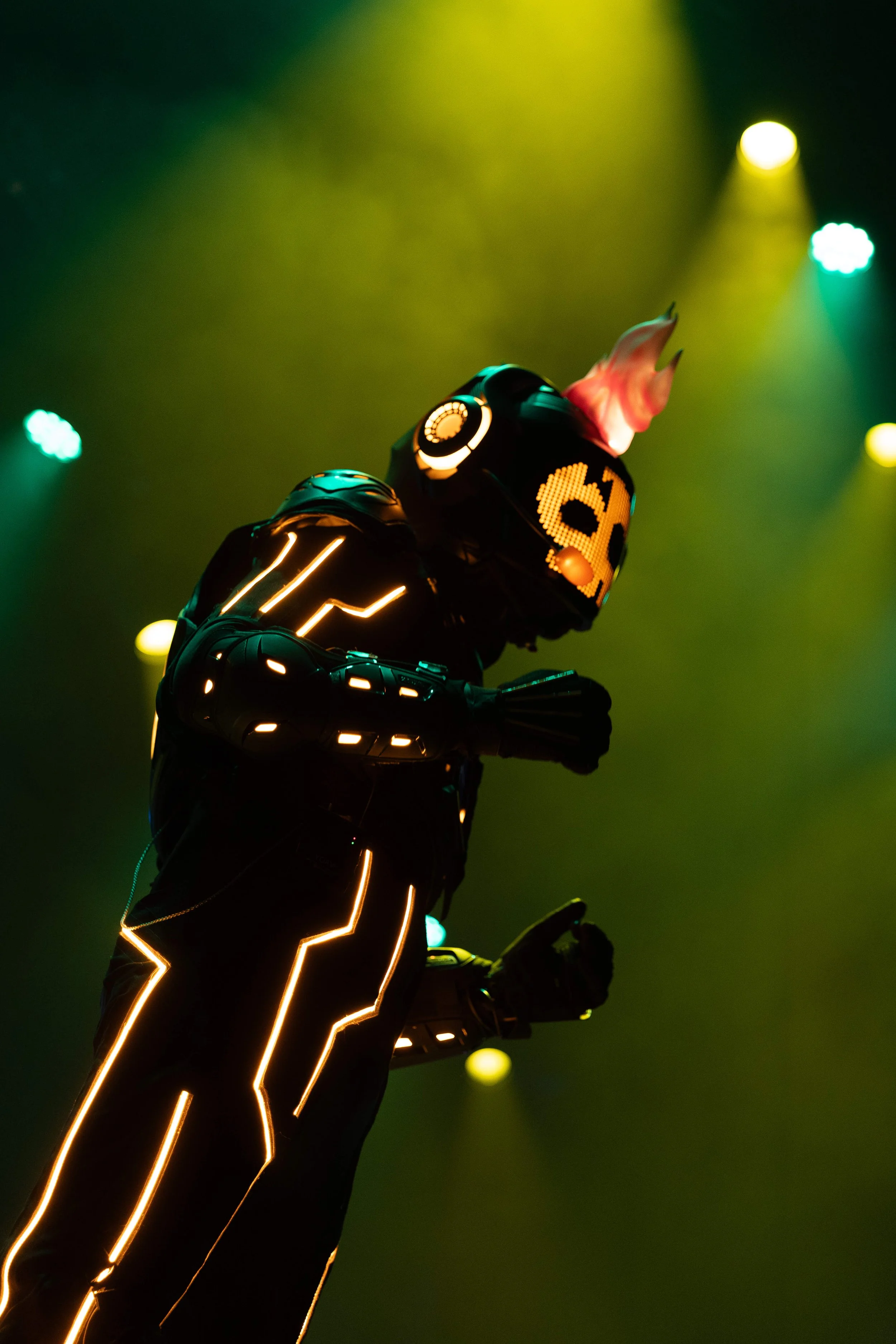 A futuristic robotic DJ with LED lighting, wearing a mask with a digital face, performing in front of stage lights with yellow, green, and cyan colors.
