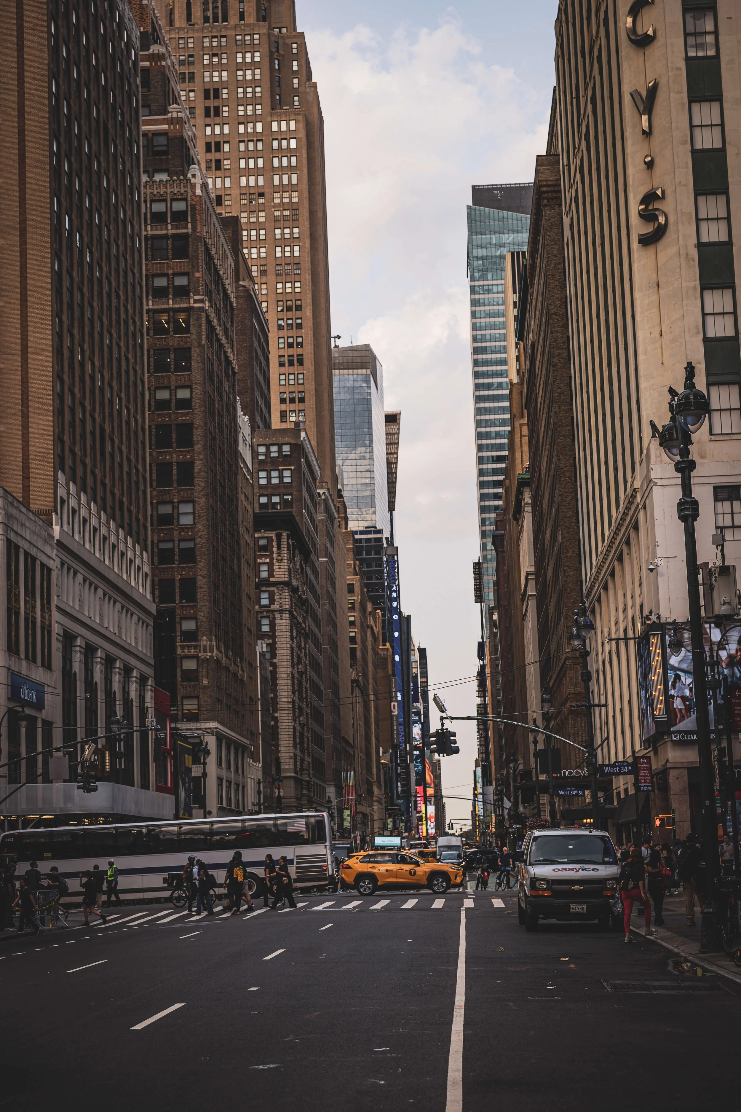 A busy city street with tall skyscrapers on either side, a yellow taxi cab, a white bus, and pedestrians crossing the crosswalk.