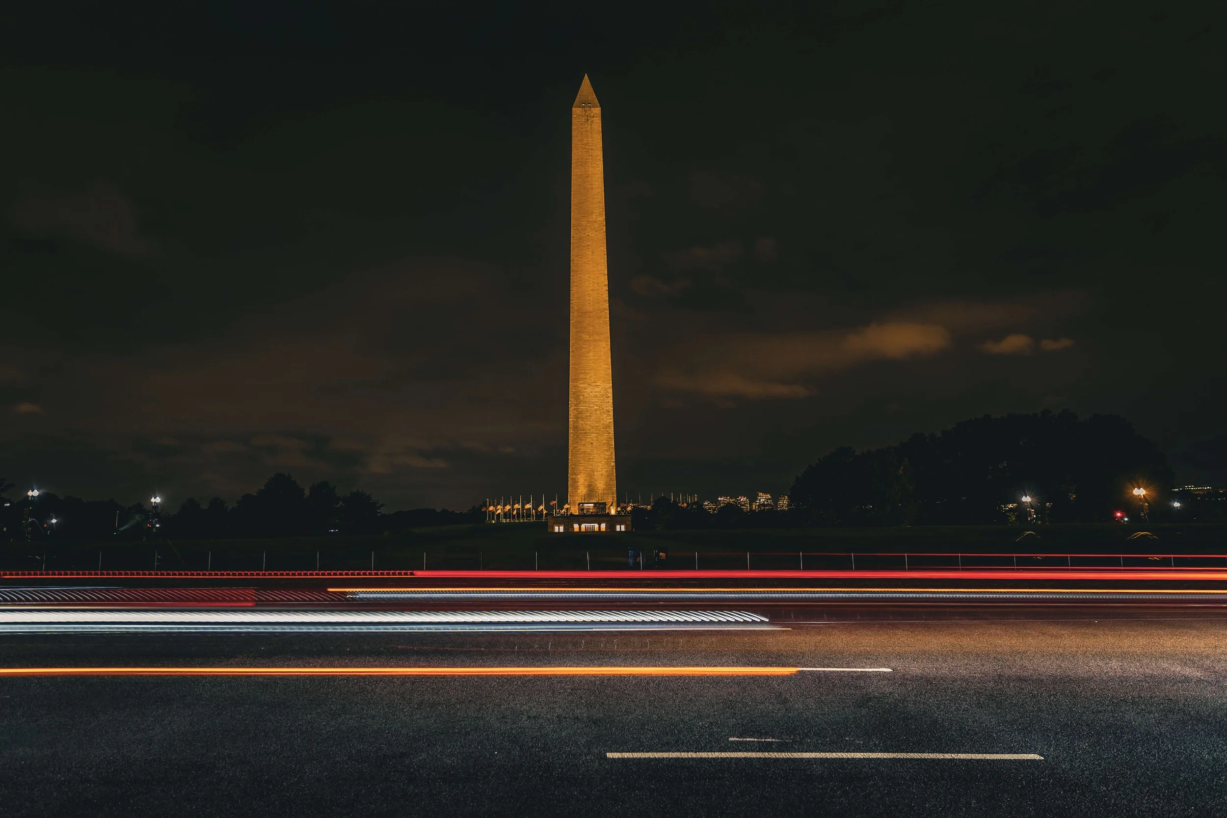 Nighttime view of the Washington Monument in Washington D.C., illuminated and standing tall in the background, with blurred car light trails on a highway in the foreground.