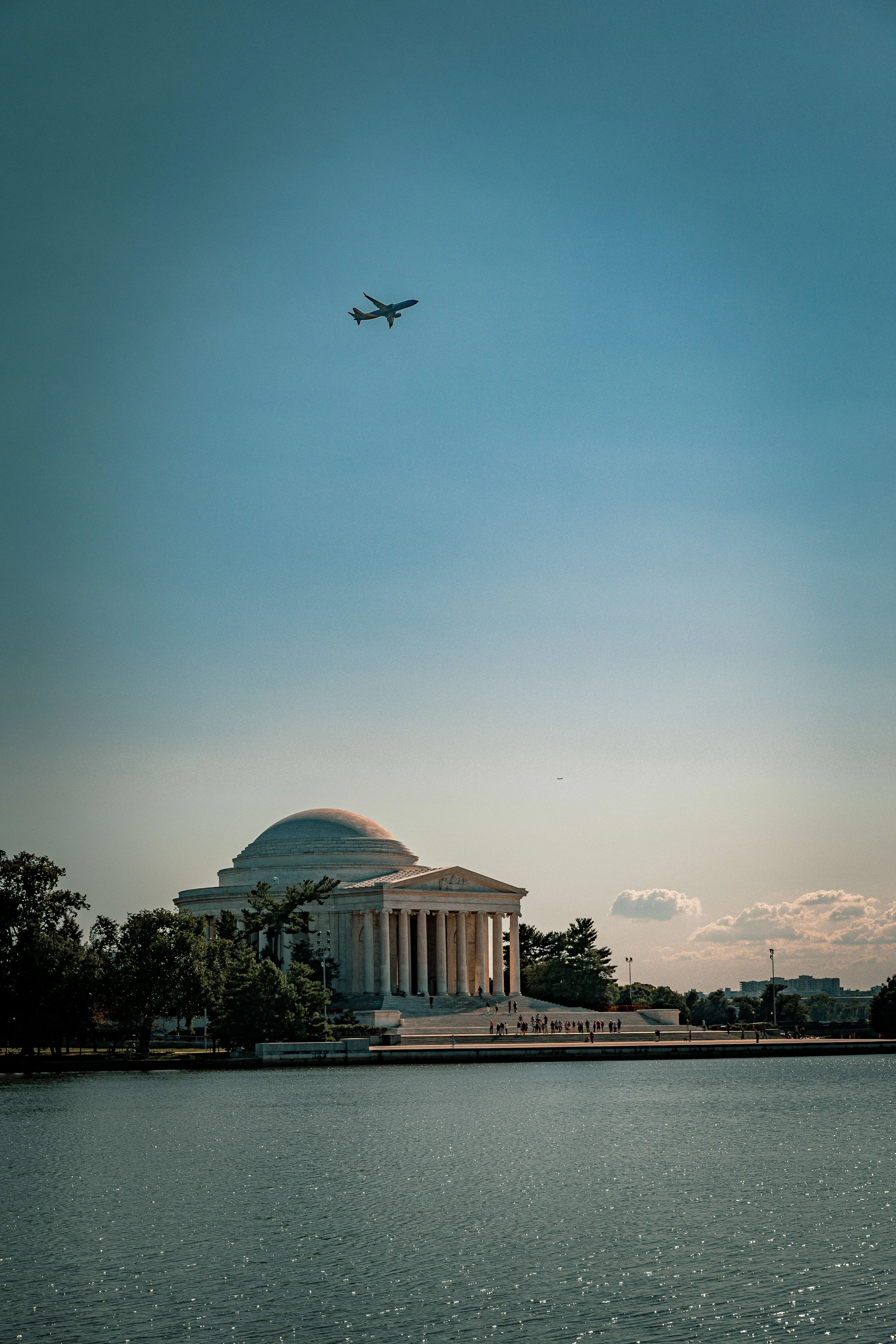 The Jefferson Memorial with an airplane flying overhead in the sky near clouds and trees reflected in the water.