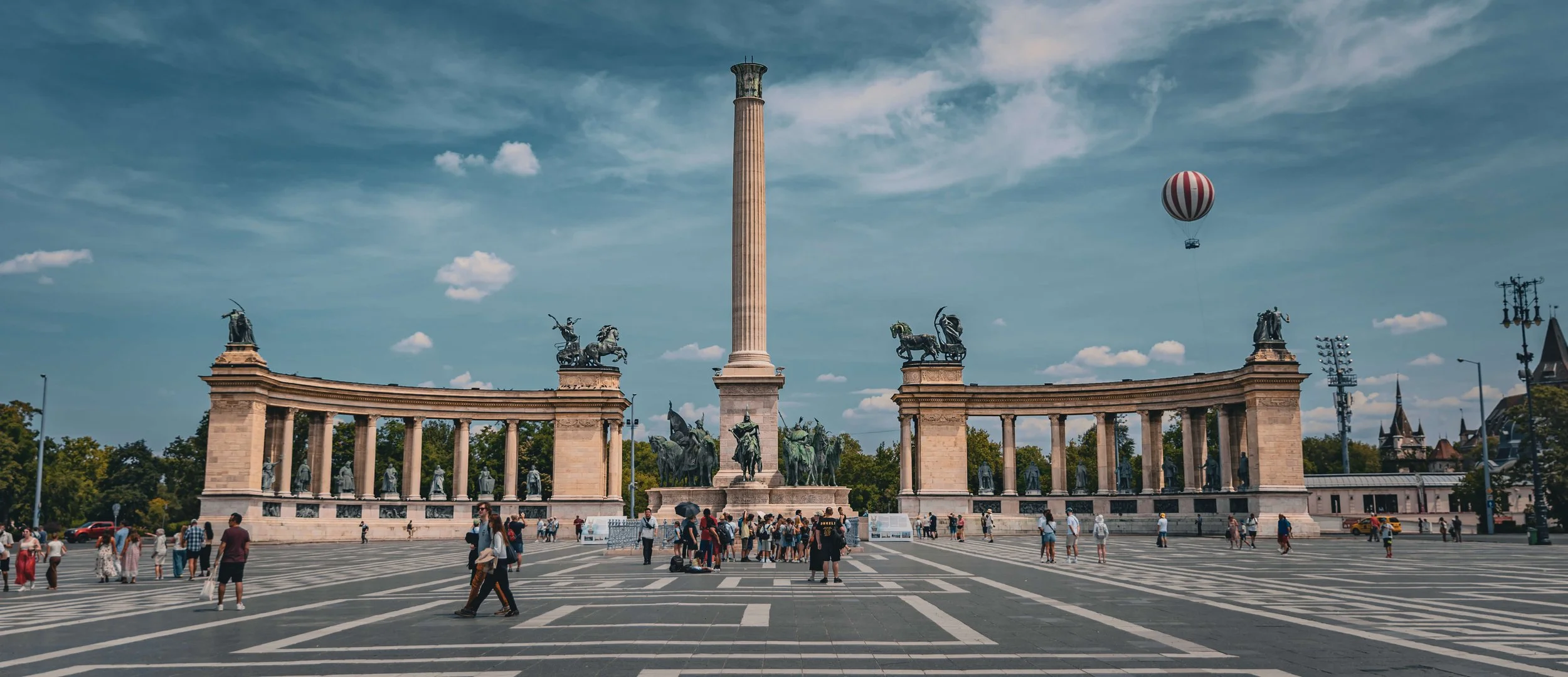 A large public square with a central monument featuring a tall column and multiple statues of horses and riders. People are walking around the square, with some standing and taking pictures. There's a hot air balloon in the sky and a background of tr