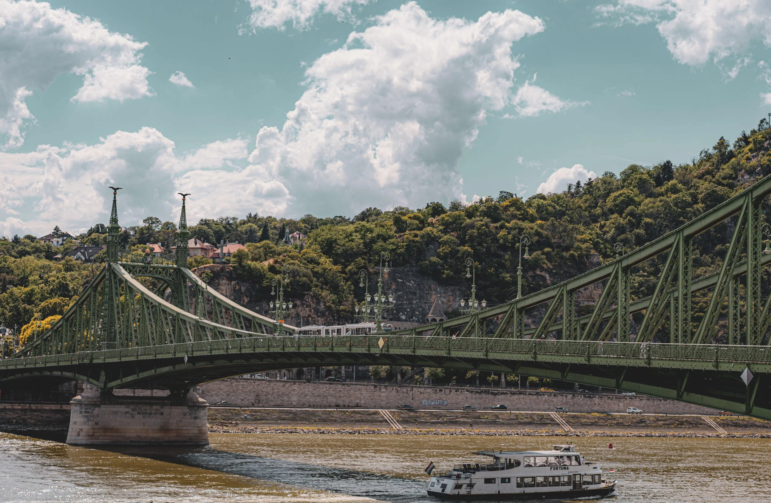 A green bridge crossing a river with a boat on the water. In the background, a hillside covered with trees and houses, with a partly cloudy sky overhead.