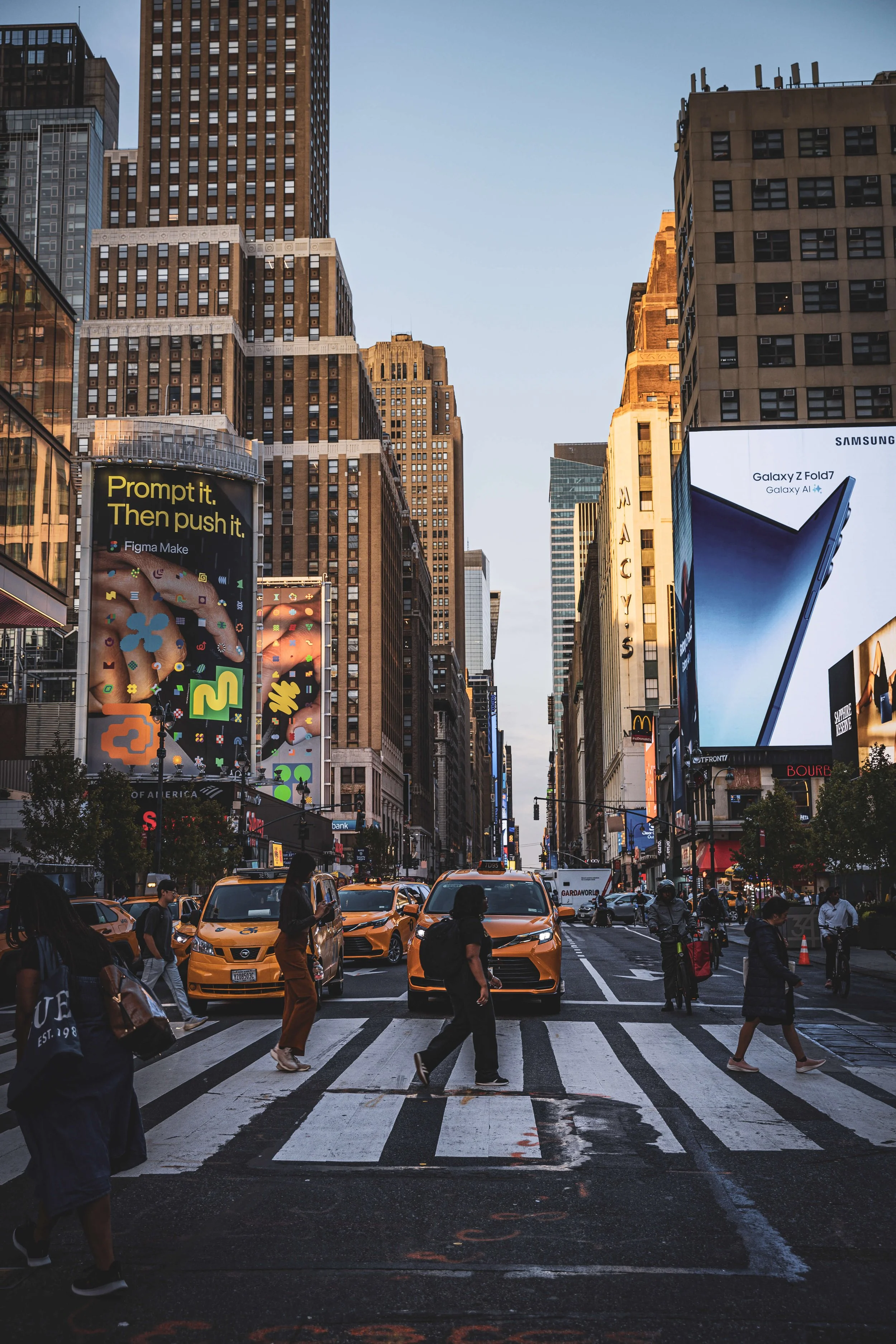 A city crosswalk at sunset in Times Square, NYC, with pedestrians, yellow taxis, and billboards advertising electronics and brands.
