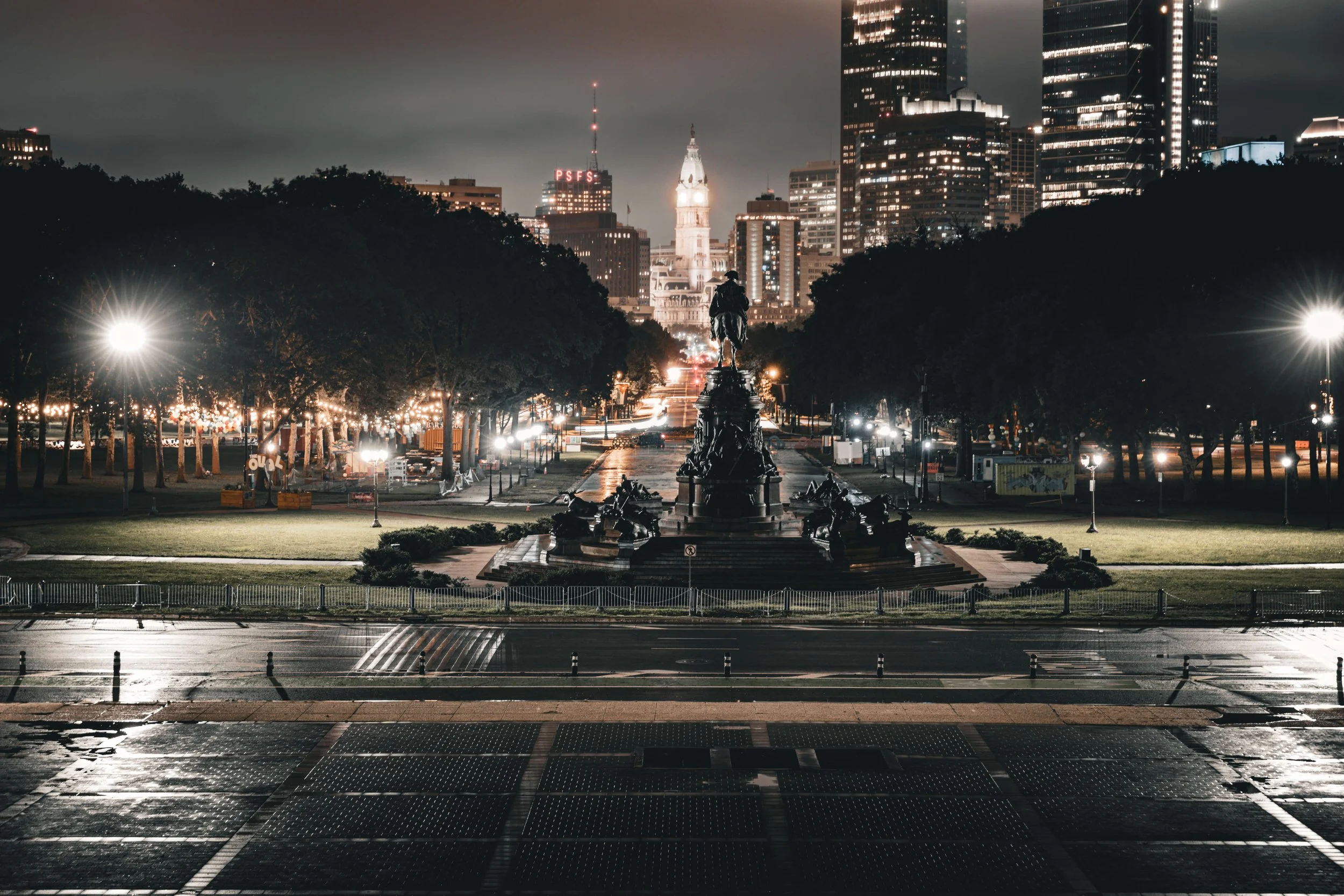 Nighttime cityscape with a central monument featuring an equestrian statue in a park, surrounded by trees, illuminated by streetlights, with tall modern buildings in the background and a clock tower in the distance.
