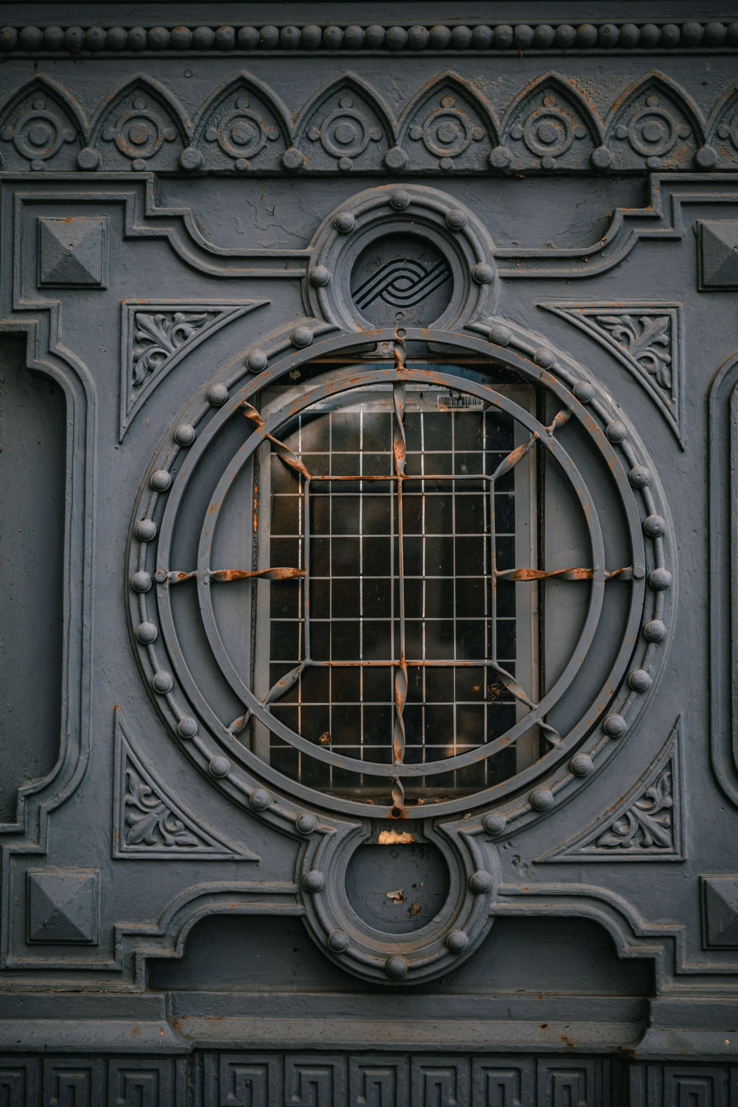 Close-up of an ornate, vintage, dark gray metal door with decorative circular and geometric patterns, a small round window with a grid, and signs of rust and weathering.