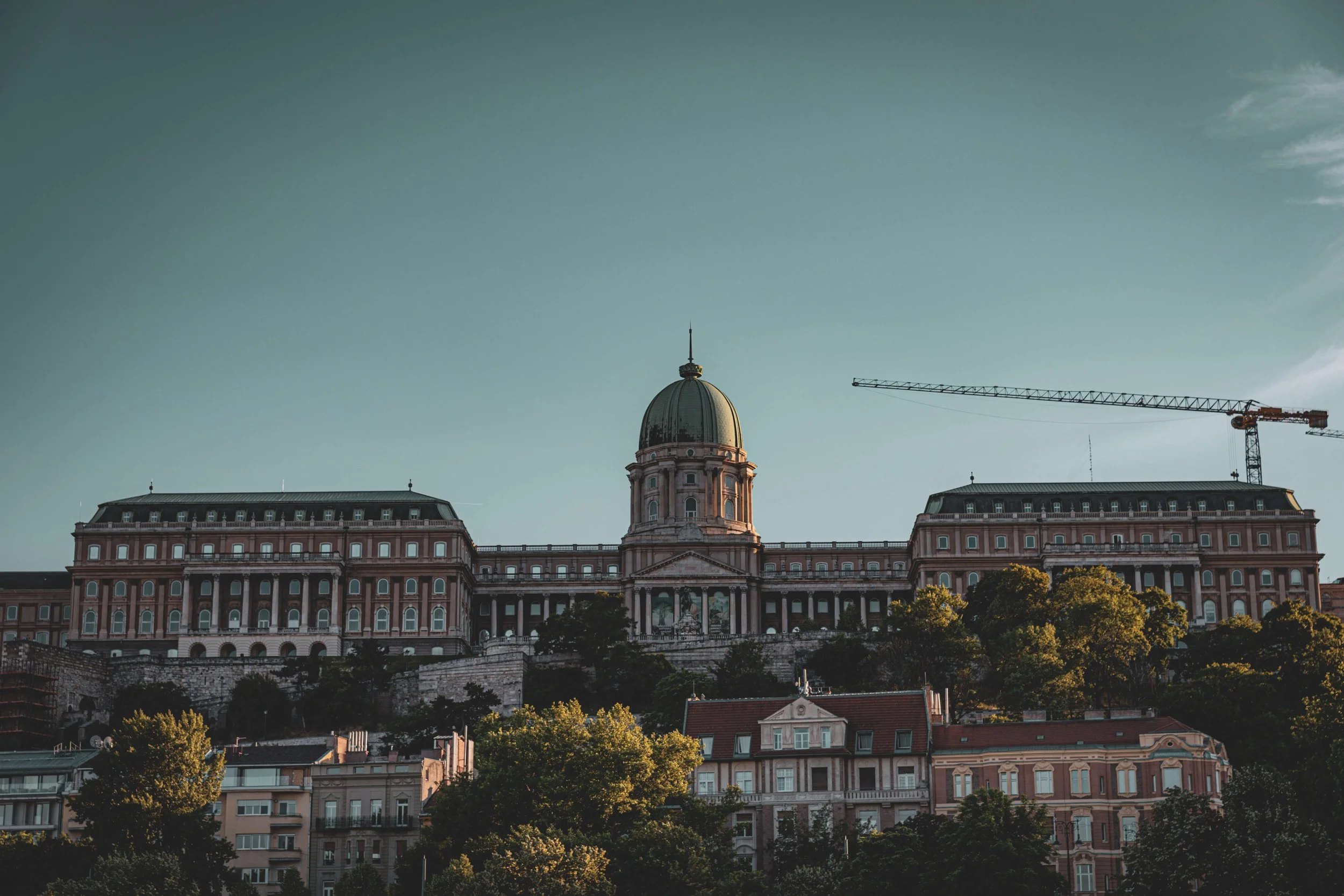 A historic building with a green dome on top, situated on a hill, with a construction crane to the right and leafy trees in front.