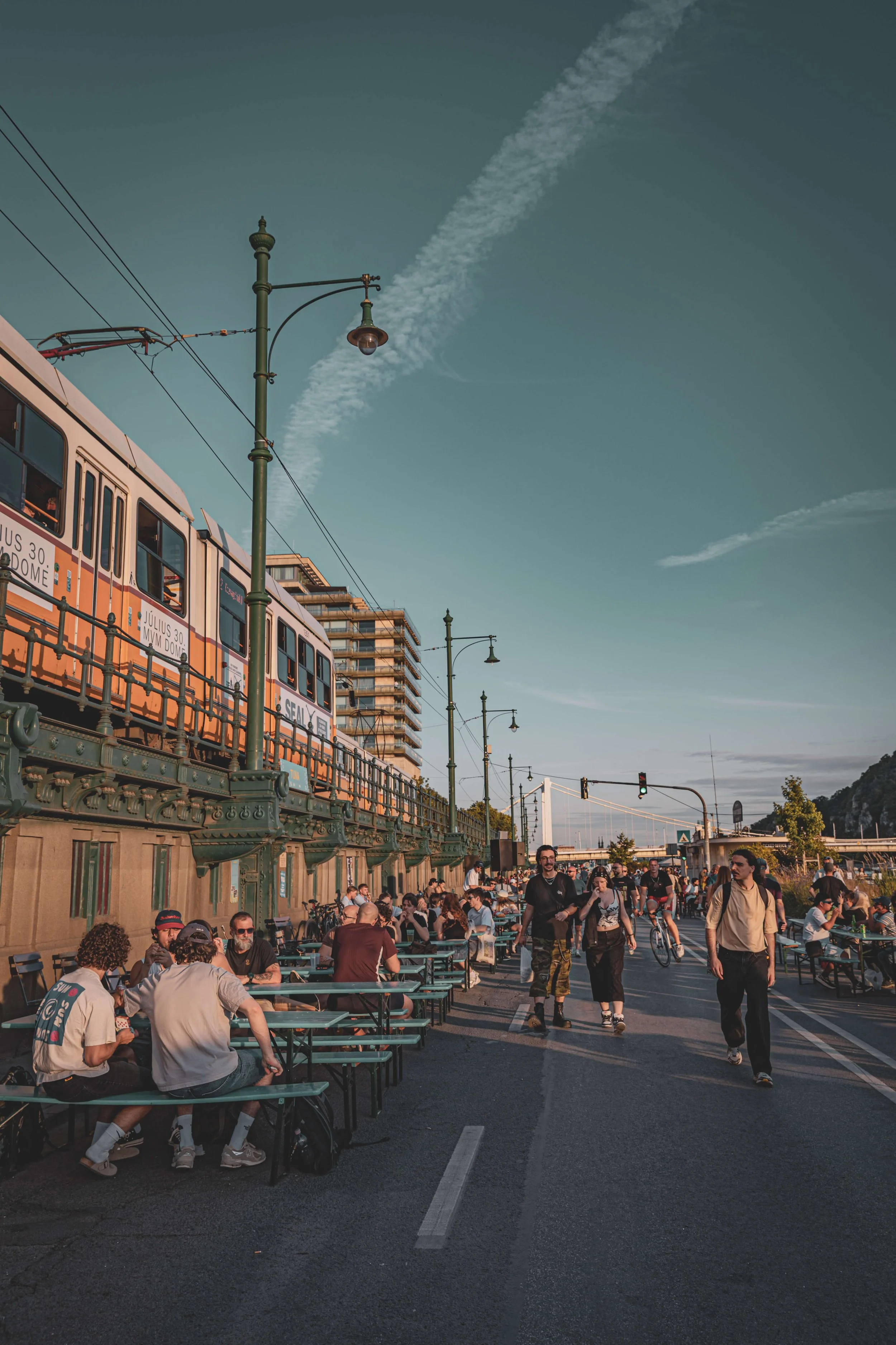 Crowd of people sitting at outdoor tables and walking near a green bridge with street lamps, trains, and buildings in the background during the evening.