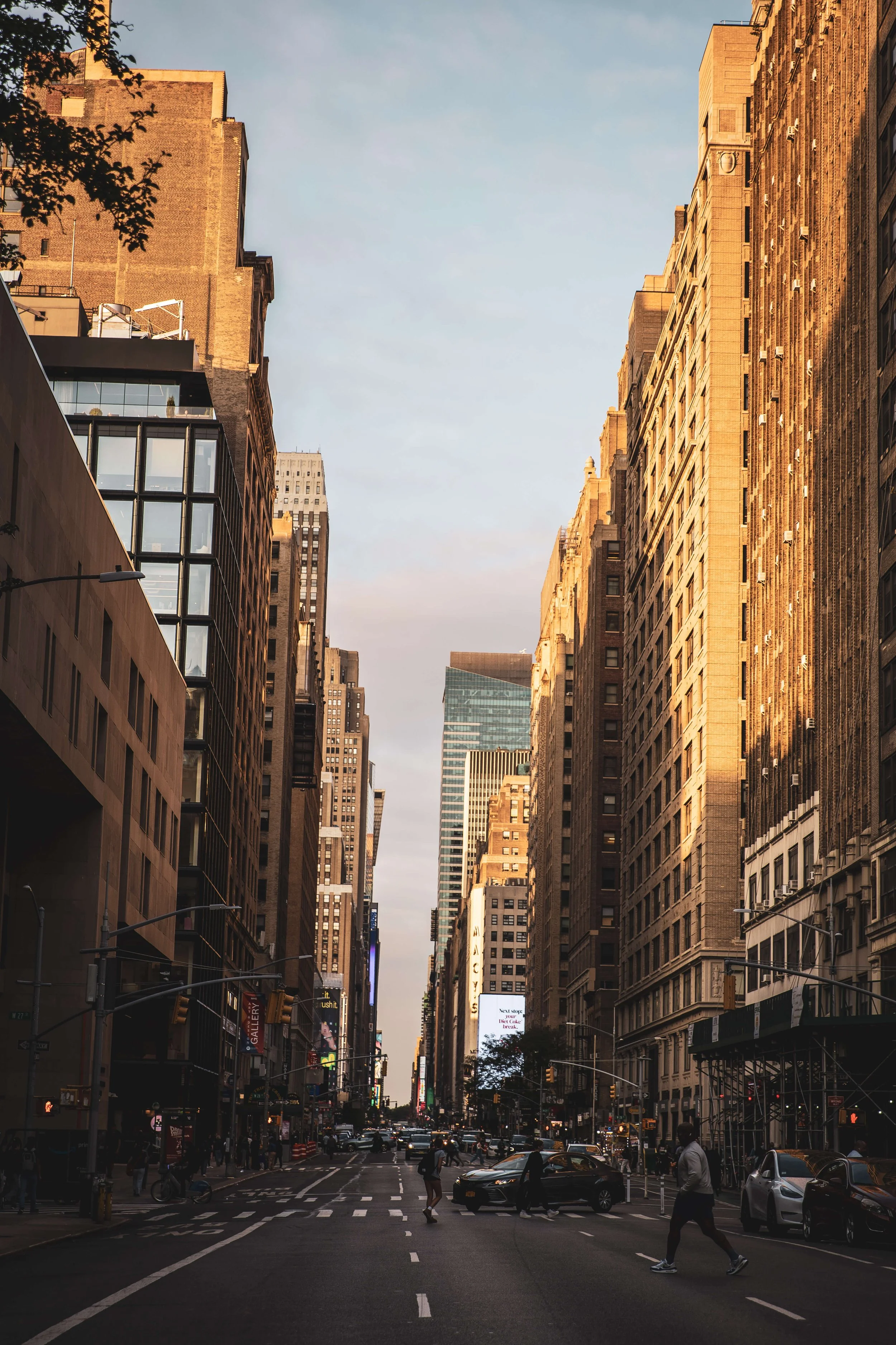 A city street scene in New York City featuring tall skyscrapers, some with reflective glass windows, with pedestrians crossing and cars on the road during daylight.