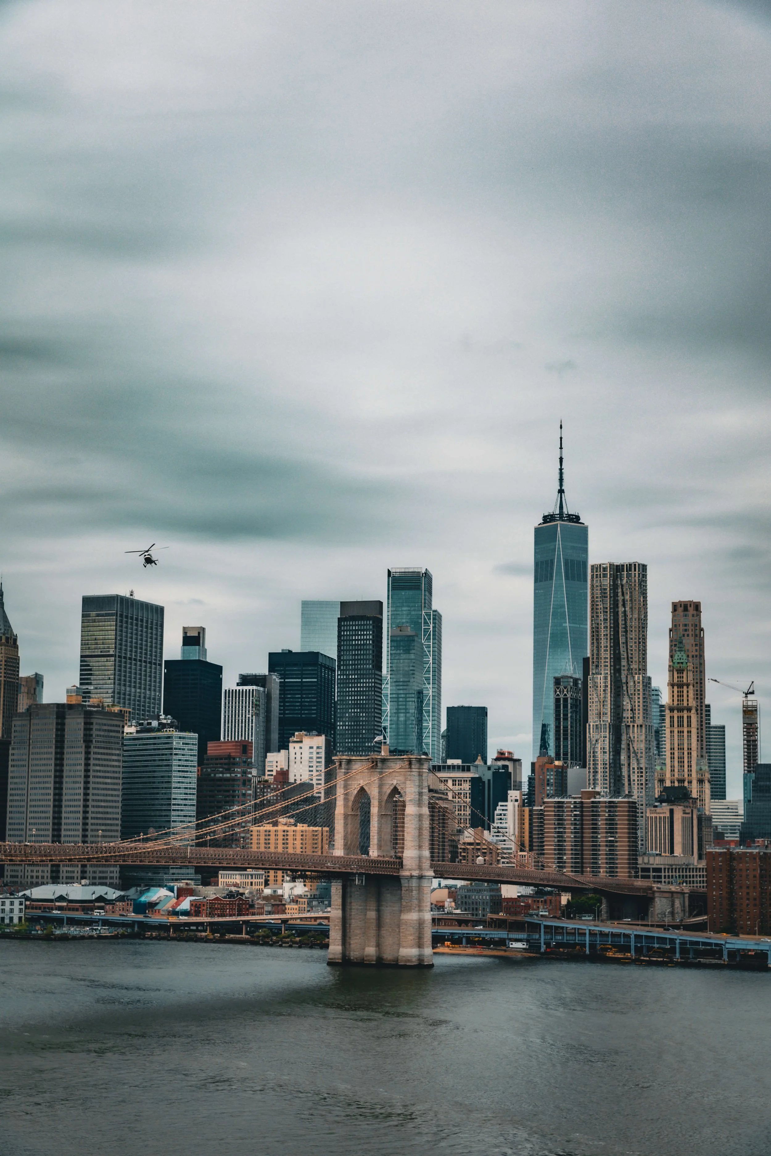 City skyline of New York City with the Brooklyn Bridge over the water and a helicopter flying in the cloudy sky.