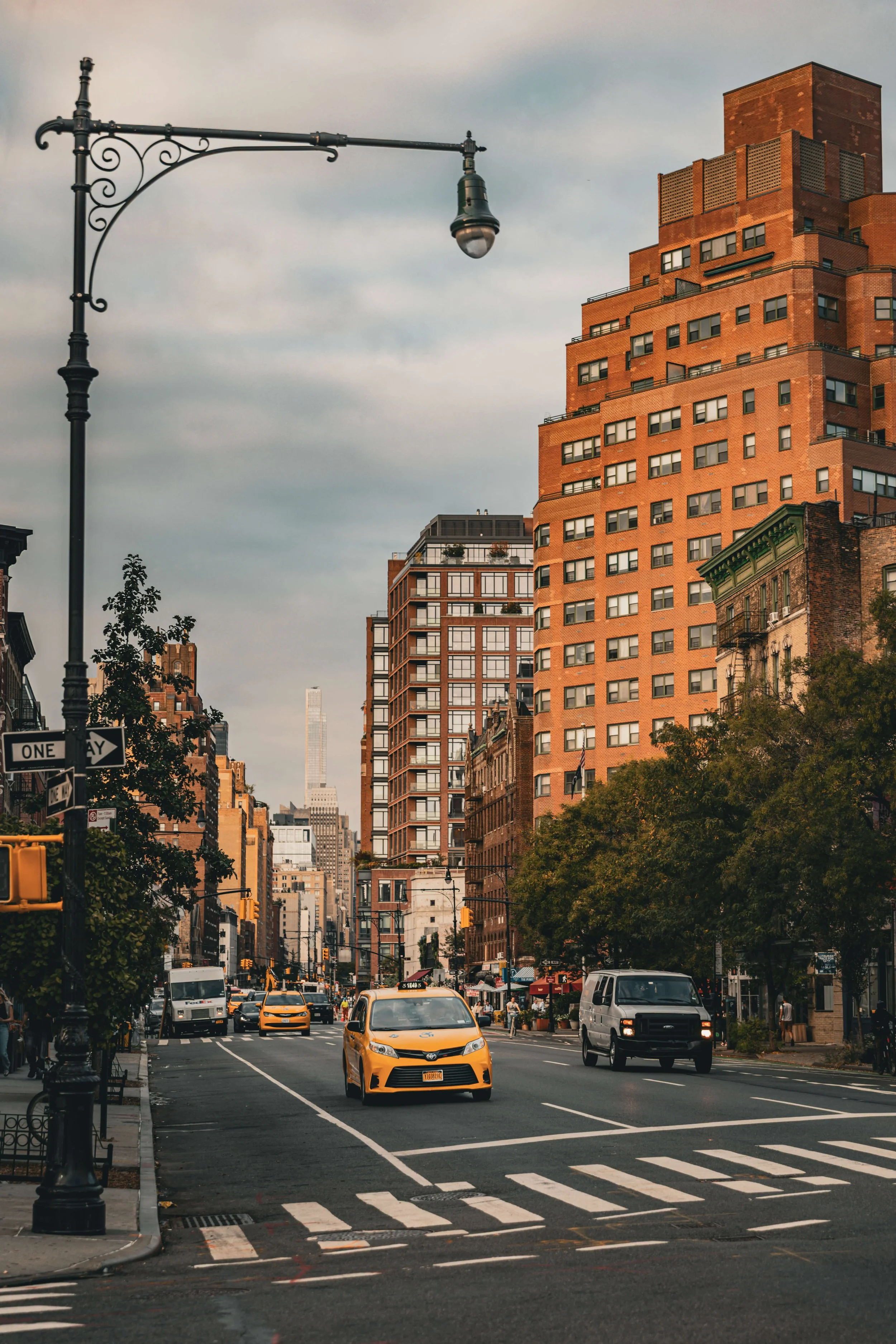 City street scene with tall buildings, yellow and white taxis, cars, pedestrians, trees, street signs, and a lamppost under a cloudy sky.