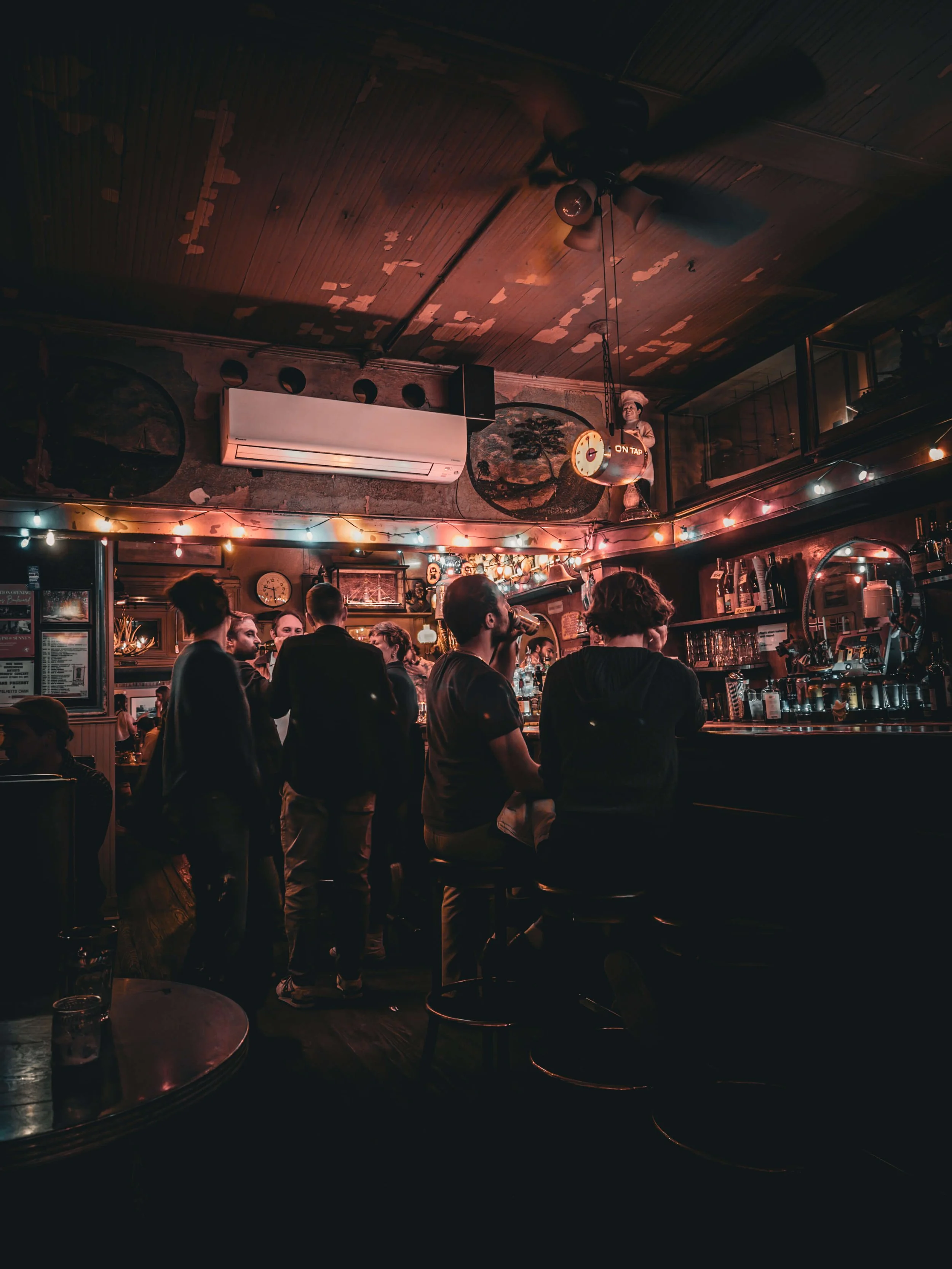 People gathered at a dimly lit bar with string lights, seated on bar stools, engaging in conversation, with bottles and glasses on the bar counter and a decorative mural on the wall.