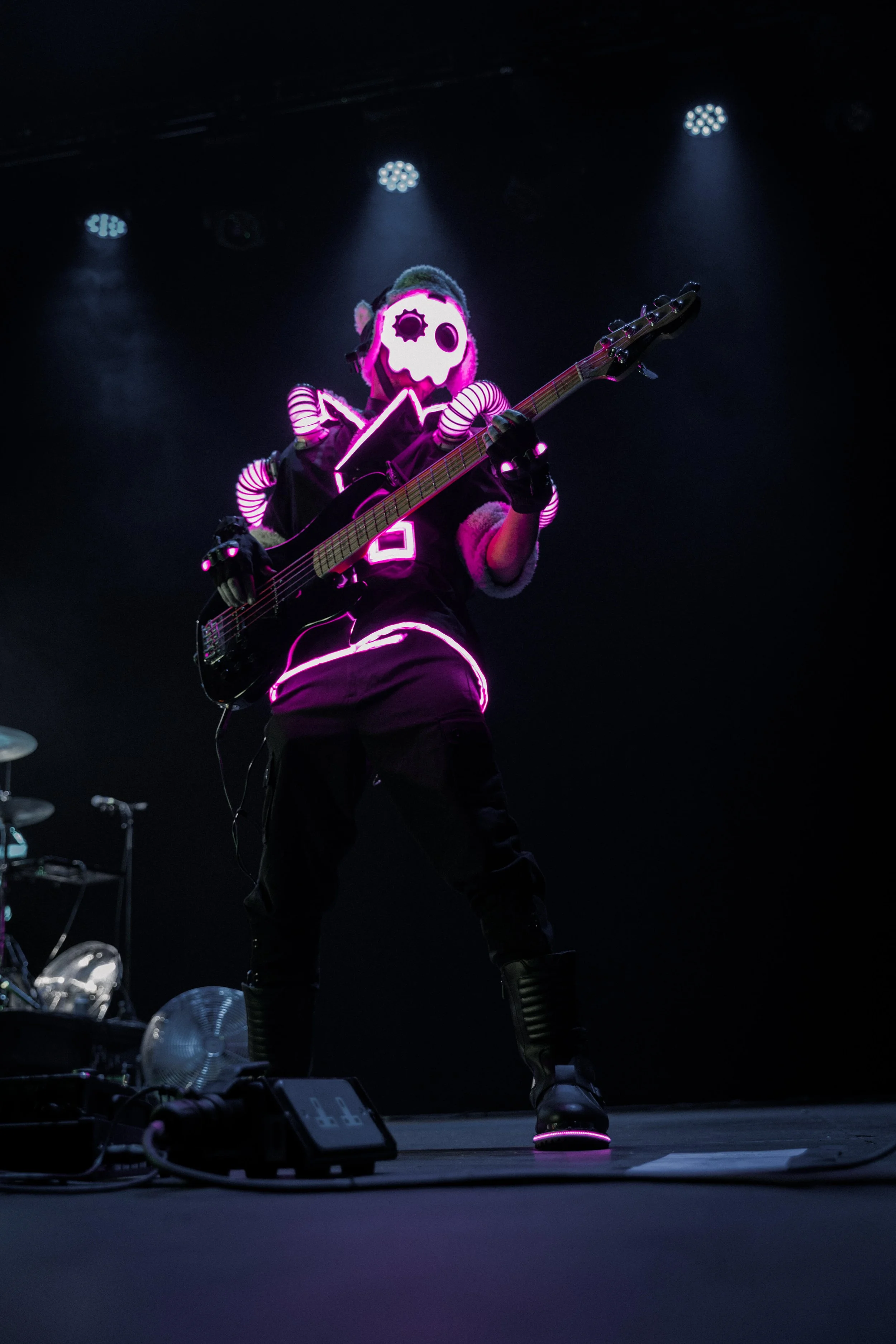 Performer on stage wearing a glowing skull mask, playing an electric bass guitar, illuminated by pink and purple neon lights in a dark setting.