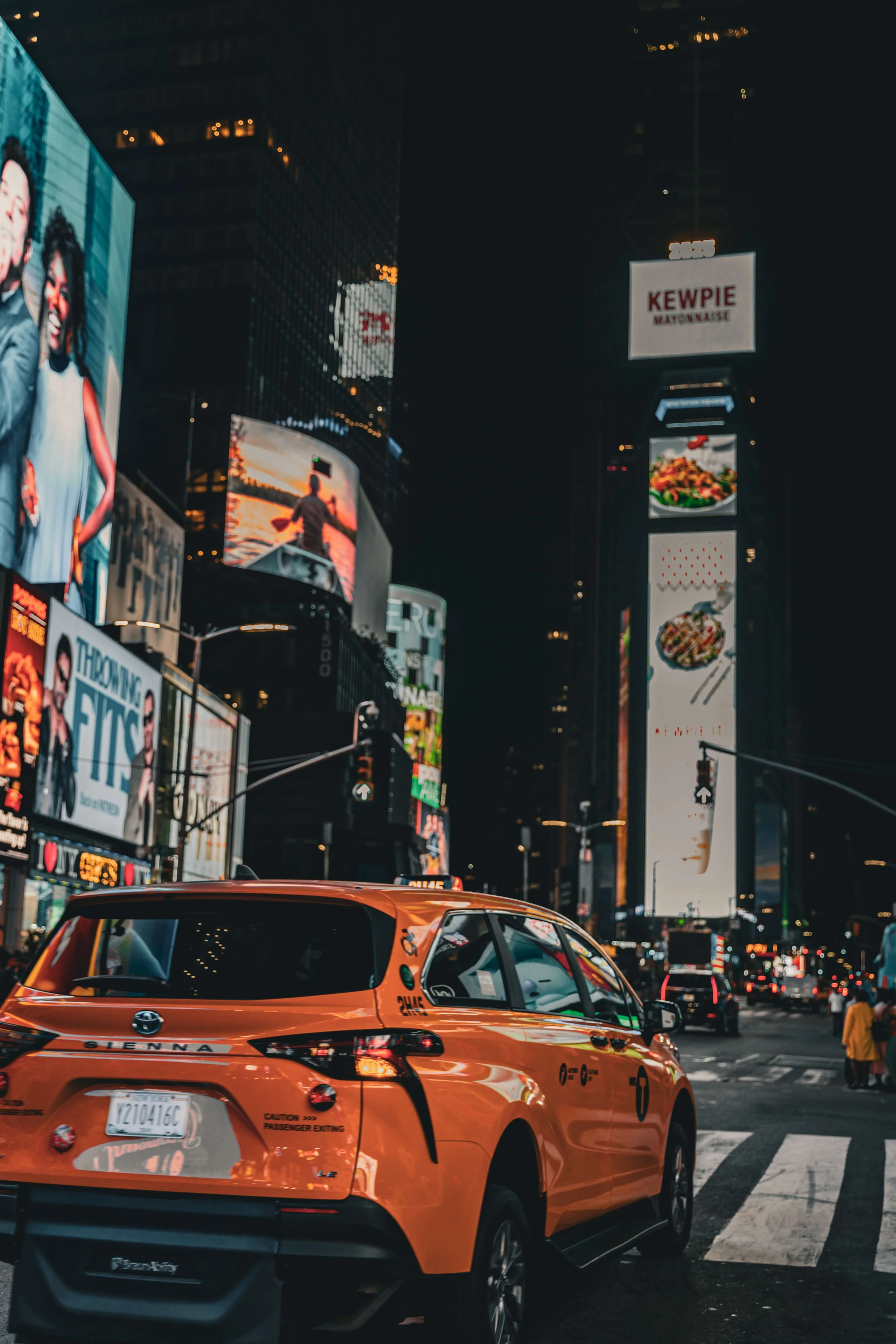 A yellow taxi cab in Times Square at night, with brightly lit billboards and digital screens in the background.