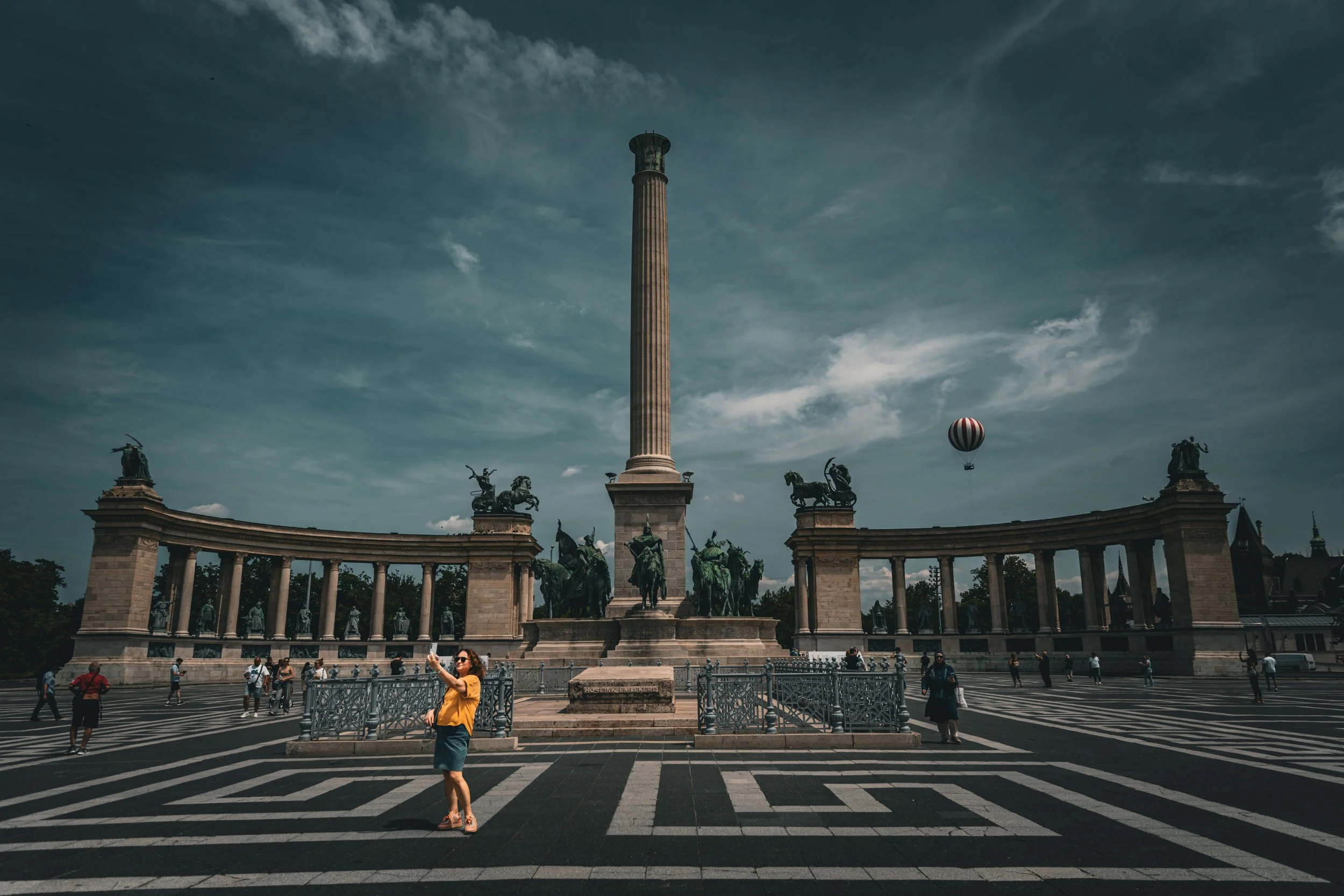 Large monument with a tall column and statues at base, crowds of people in front, and a hot air balloon in the sky.