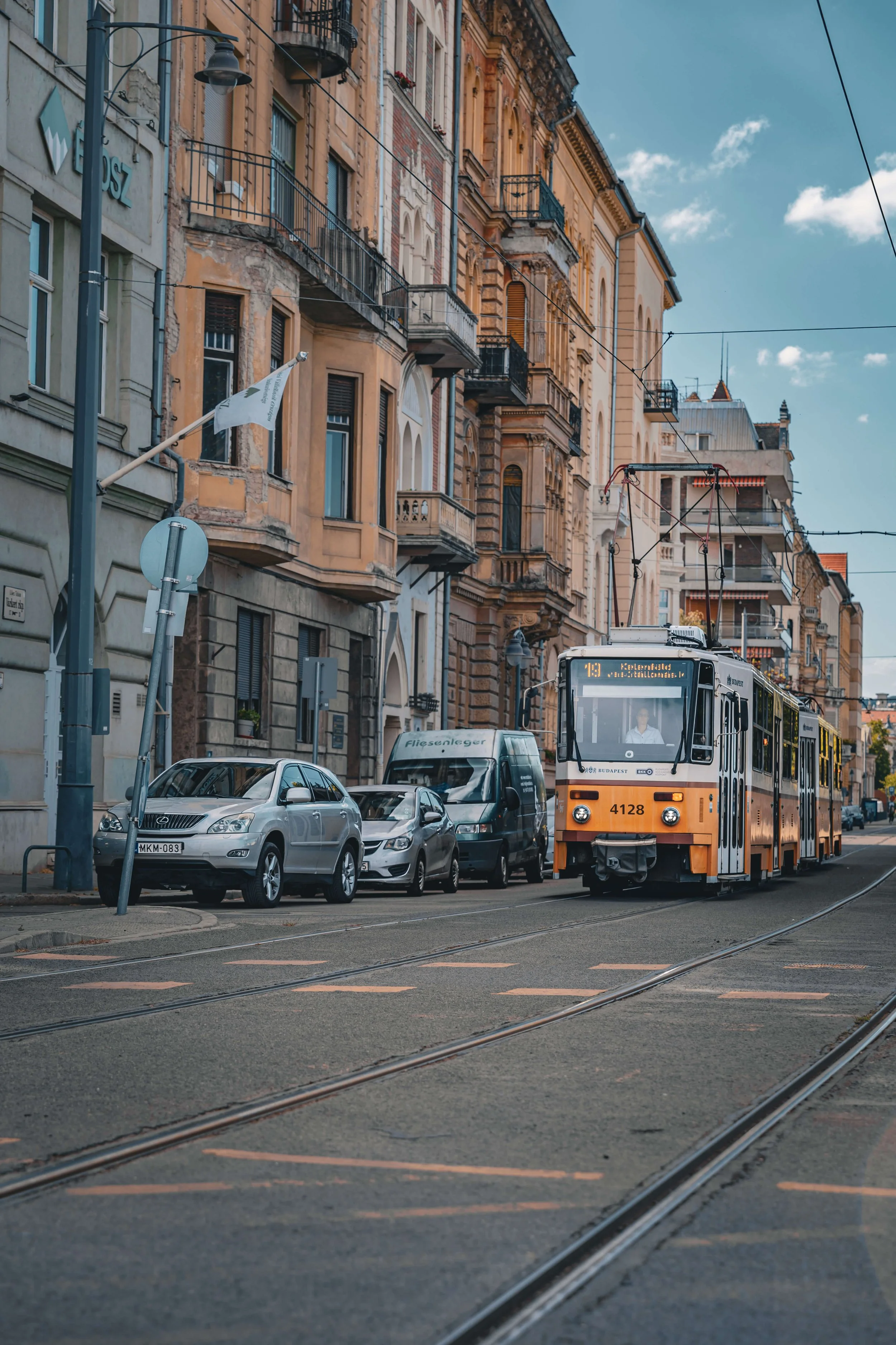 City street with parked cars and a tram traveling on tracks. Tall, old buildings with balconies and hanging wires line the street, under a partly cloudy sky.