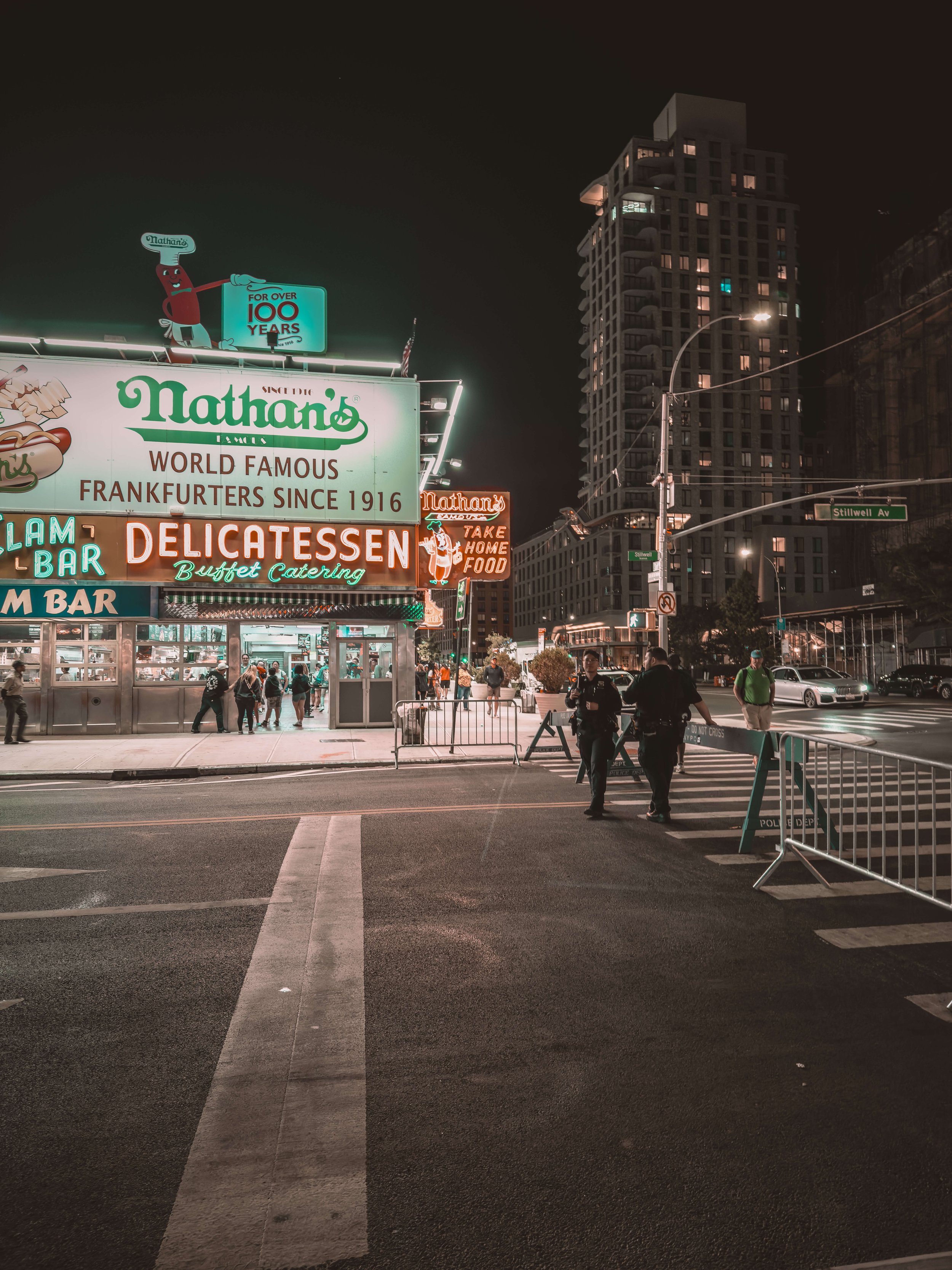 Nighttime street scene outside Nathan's Famous hot dog stand, illuminated neon signs, pedestrians and police officers near a crosswalk, tall buildings in the background.