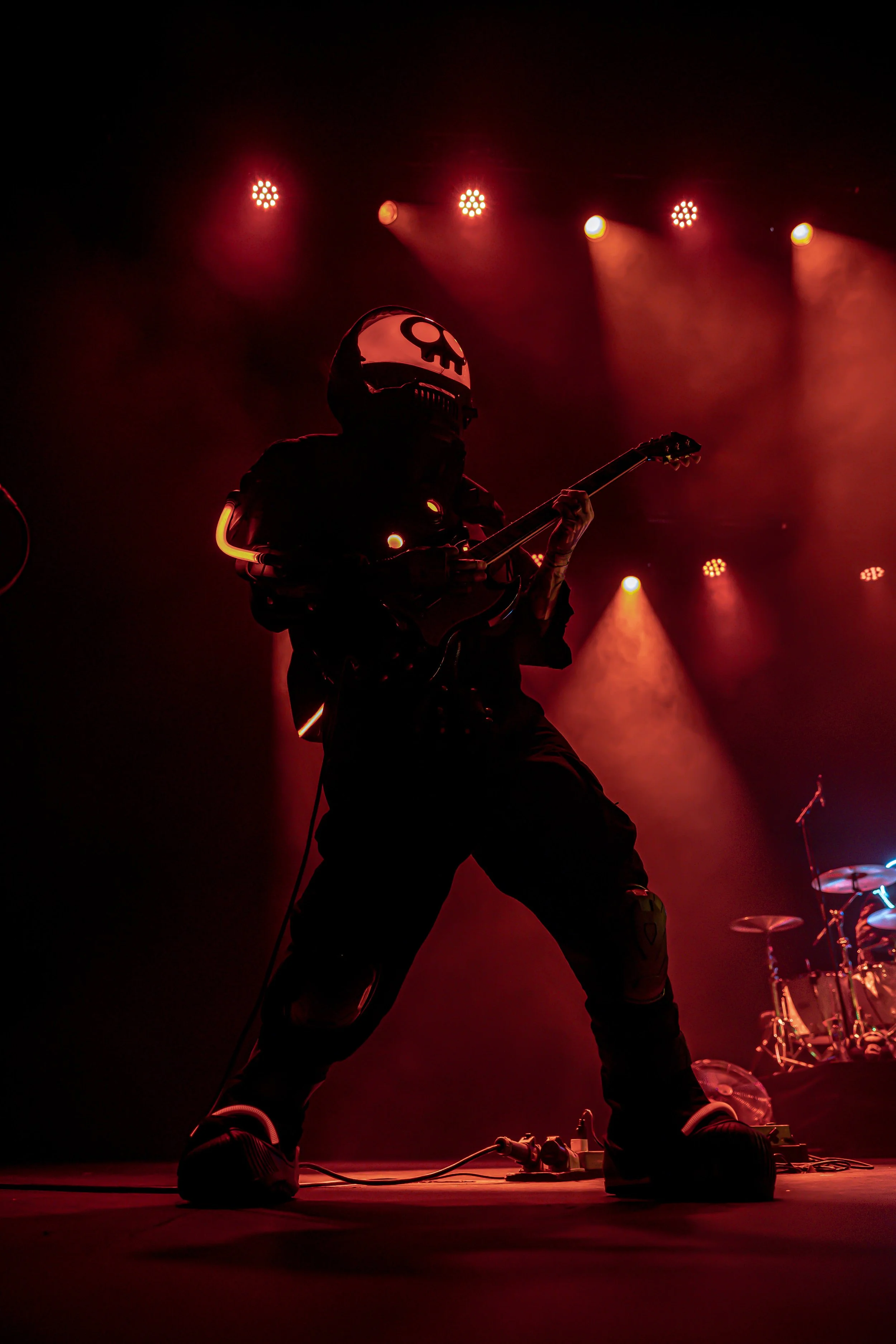 A performer wearing a skull mask and dark clothing playing an electric guitar on stage with red and orange lighting.