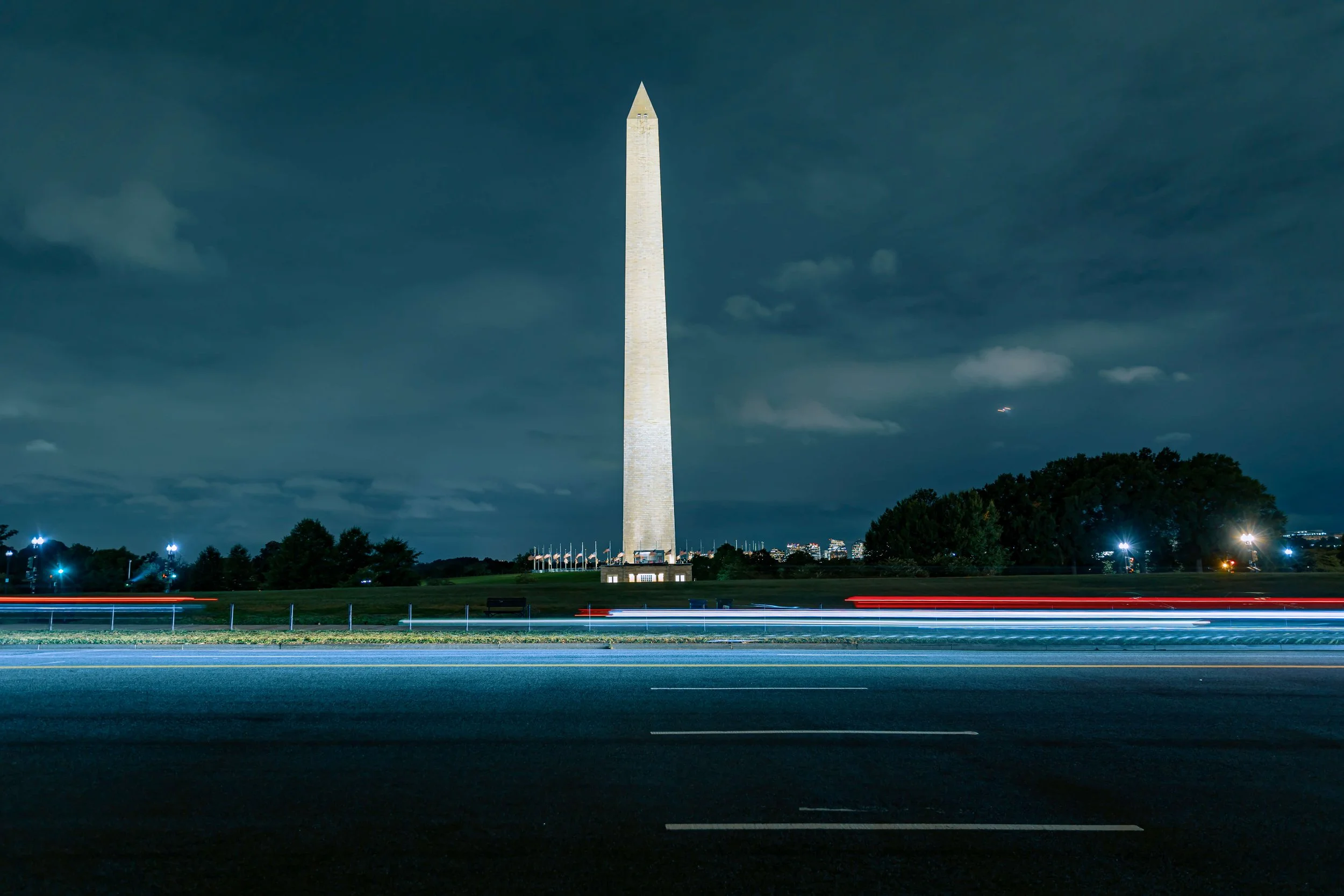 Nighttime view of the Washington Monument with light trails from passing vehicles in the foreground and a cloudy sky above.