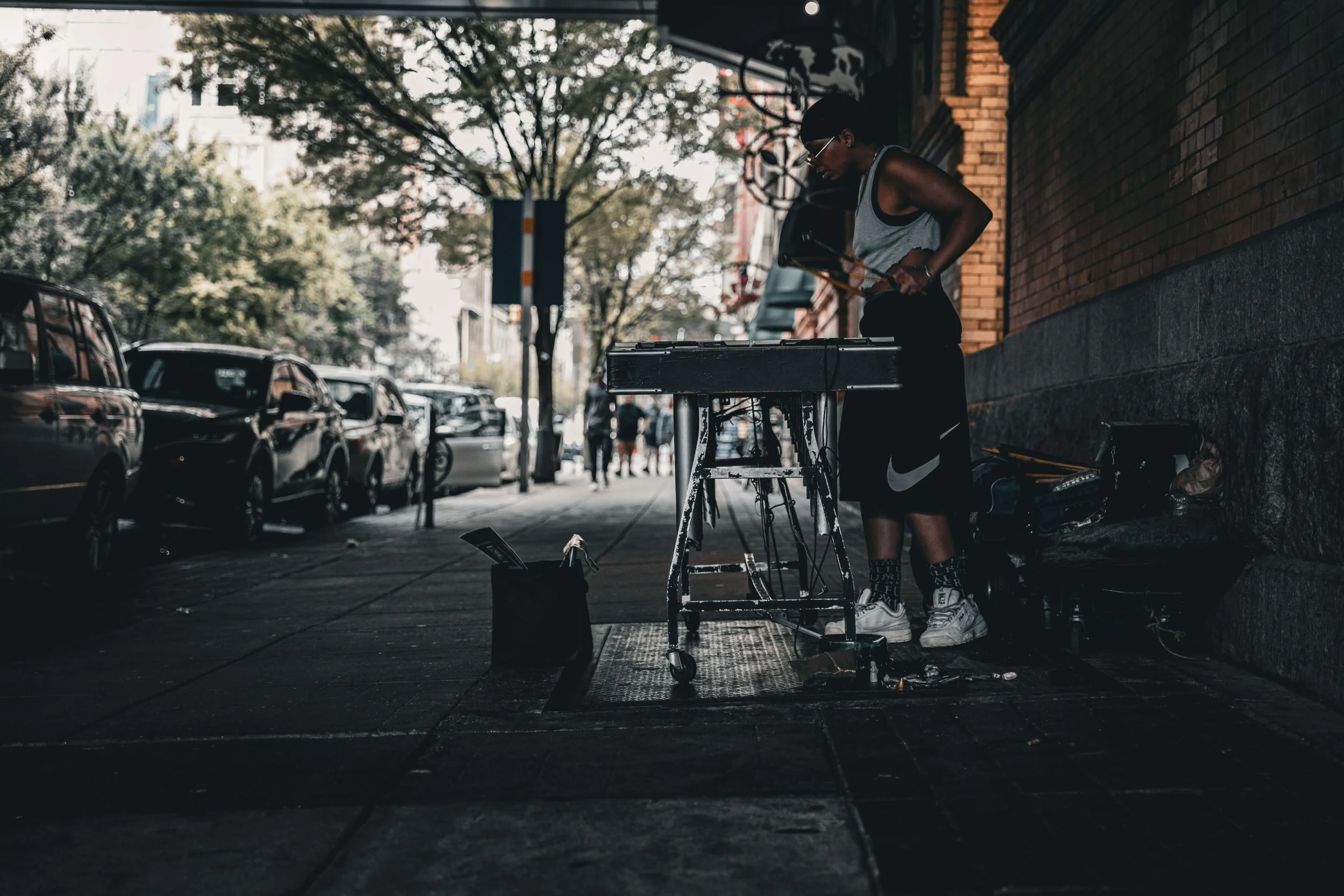 A person with sunglasses and athletic clothing performing as street musician on a sidewalk, with cars parked along the street and trees in the background.