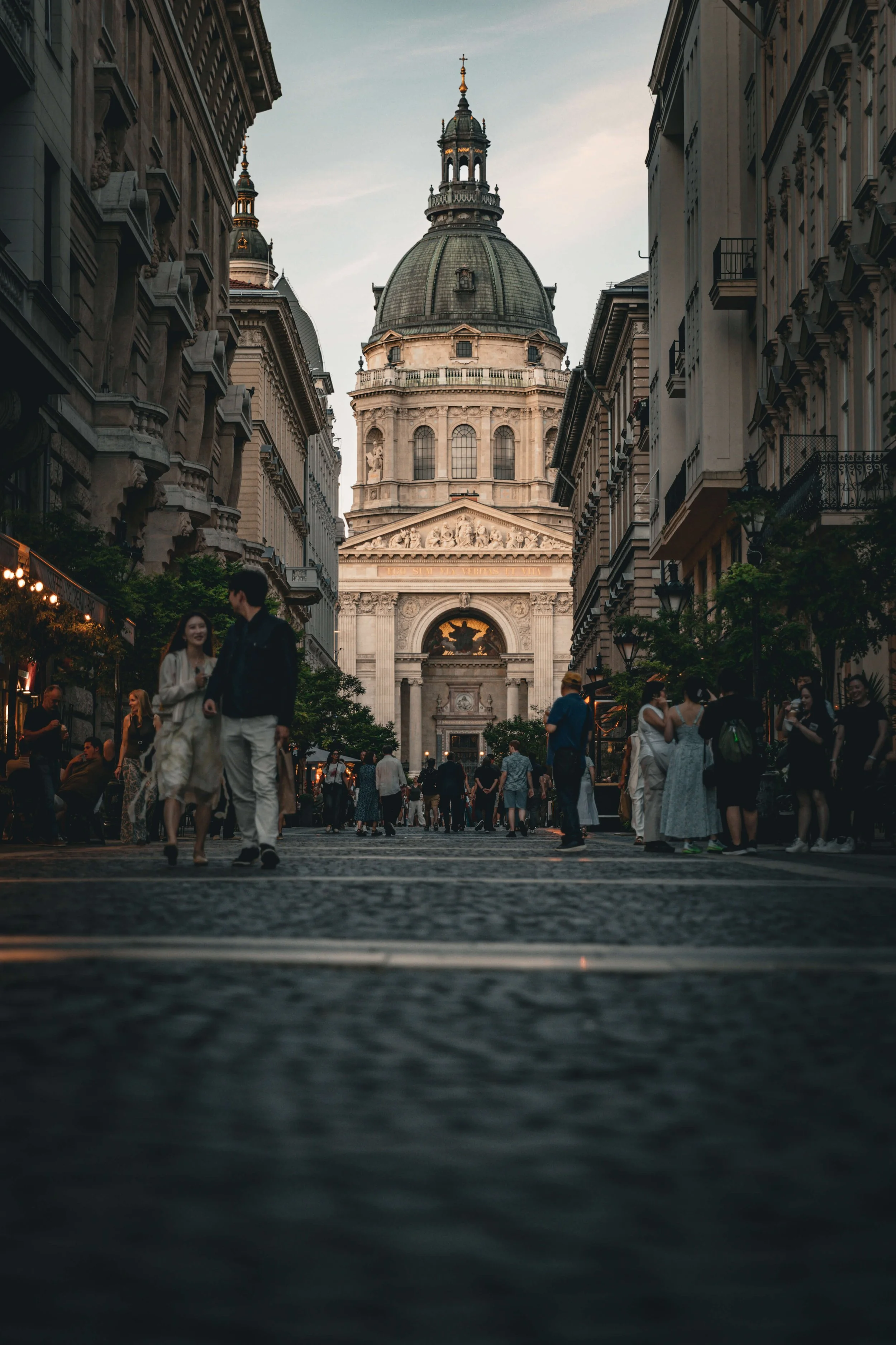 People walking on a busy street with historic buildings and a domed church in the background, during the evening.