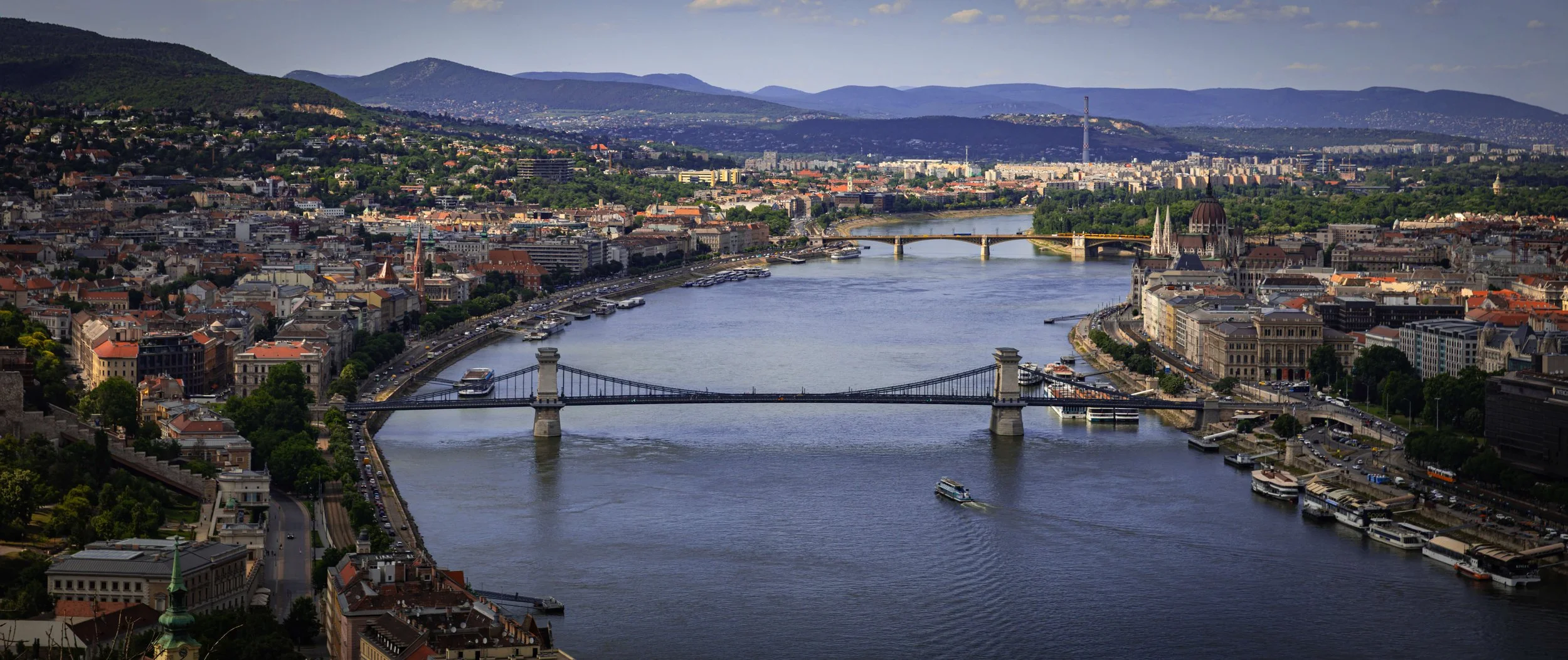 A panoramic view of a river running through a city, with several bridges, boats, and historic buildings, surrounded by green hills and mountains.